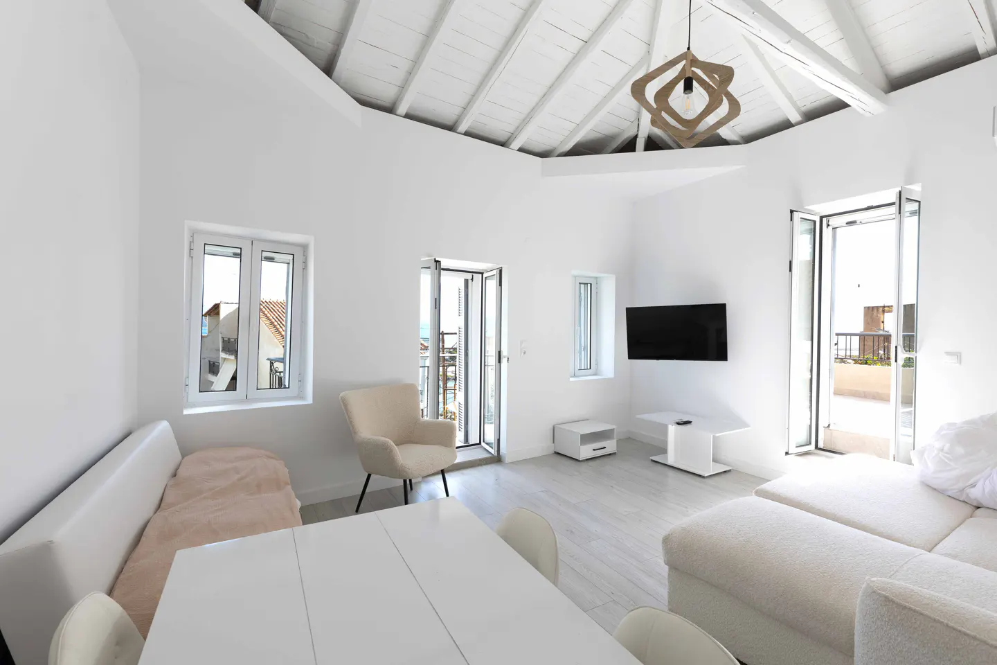 Bright, all-white living room with a dining table, sofa, chair, and TV. Windows and doors offer outdoor views. A modern light fixture hangs from the white ceiling.