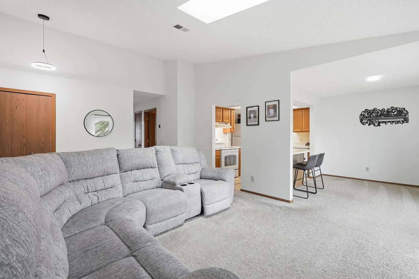 Bright living room with a gray sectional sofa, white walls, and gray carpet. A kitchen is visible in the background.
