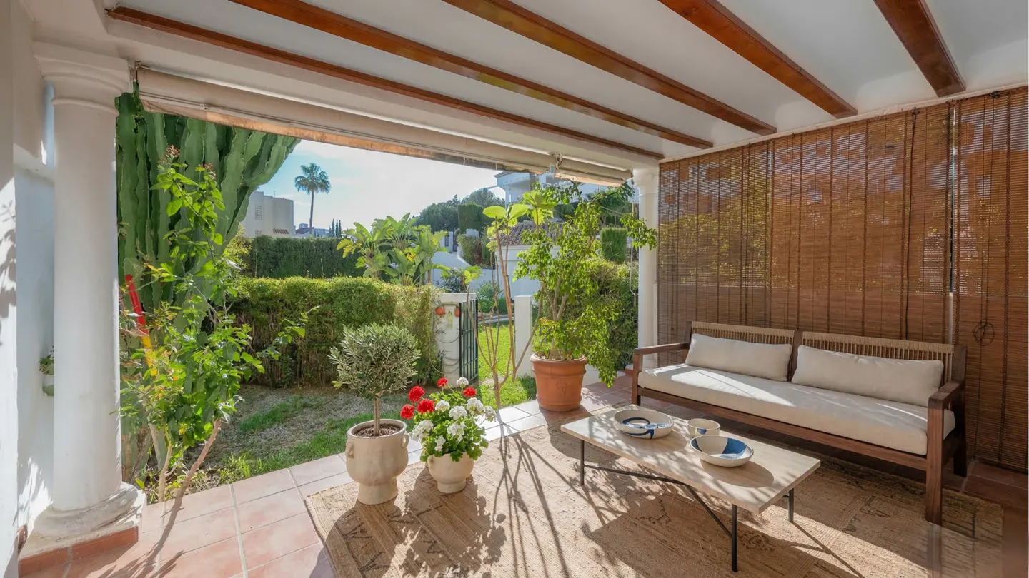 Covered patio with a wooden sofa, coffee table, and potted plants. A garden with green bushes is visible in the background.