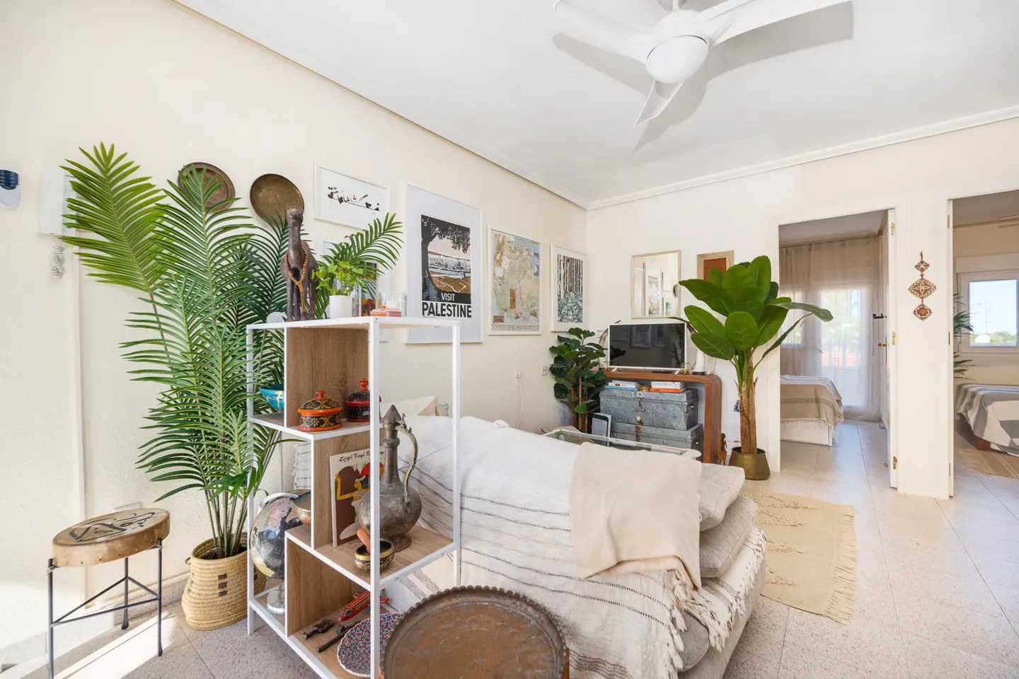 Bright living room with white walls, tiled floor, and a white ceiling fan. A sofa, shelving unit, and plants add warmth to the space. Two bedrooms are visible through doorways.