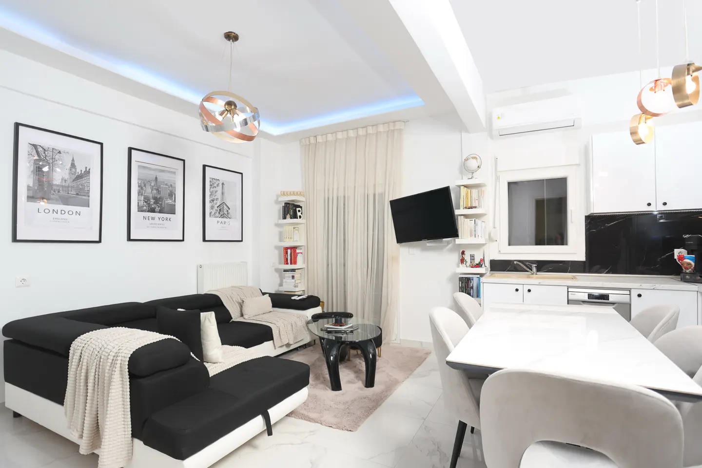 Bright, modern living room with a black and white sectional sofa, marble floors, and city-themed art. A dining table sits near a white kitchen.