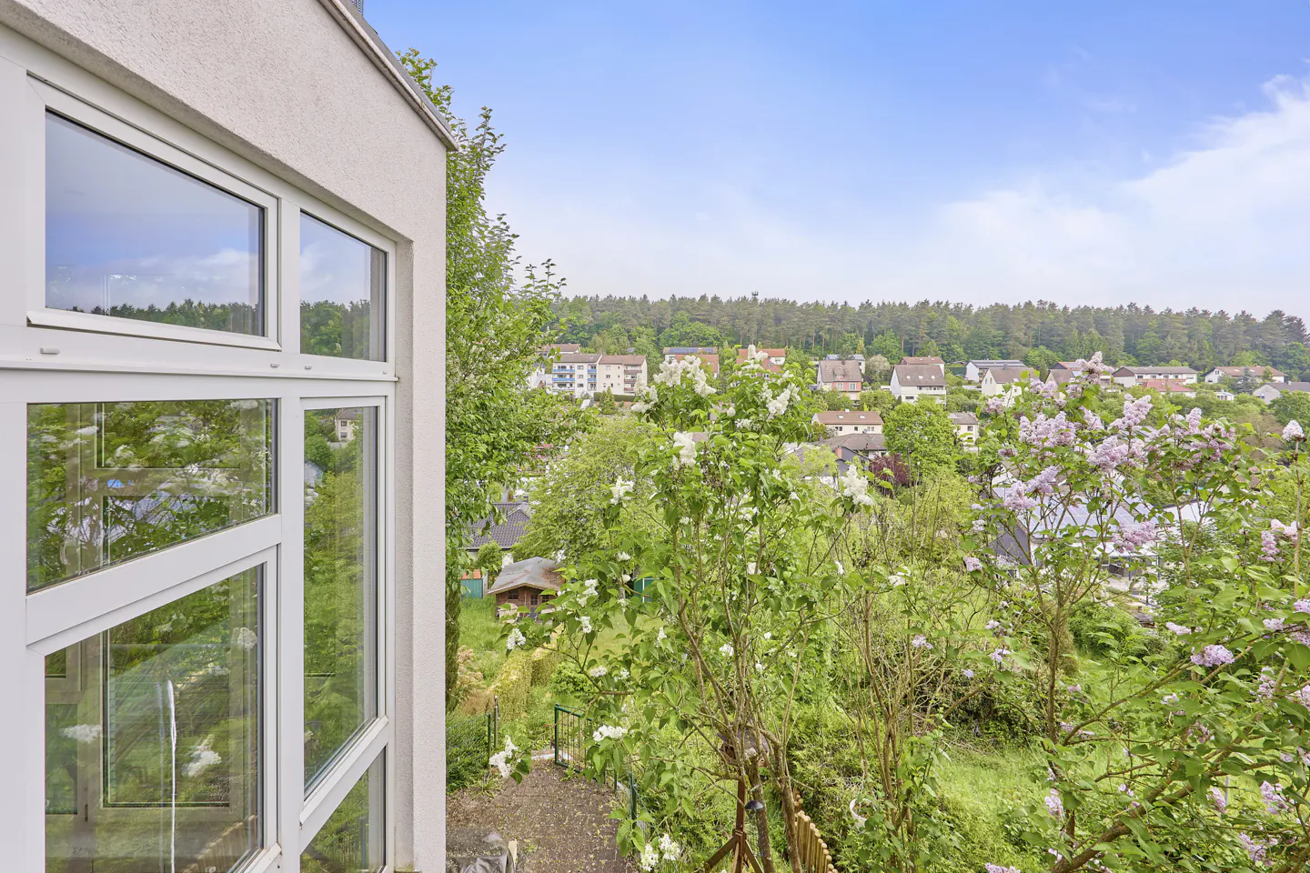 View from a window overlooking a lush green landscape with houses and trees. The sky is blue with some clouds.