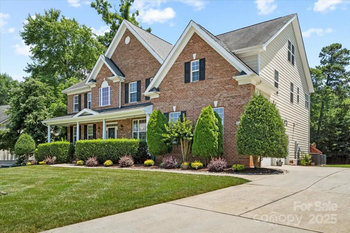 Two-story brick house with a front porch, manicured hedges, and a concrete driveway under a blue sky.