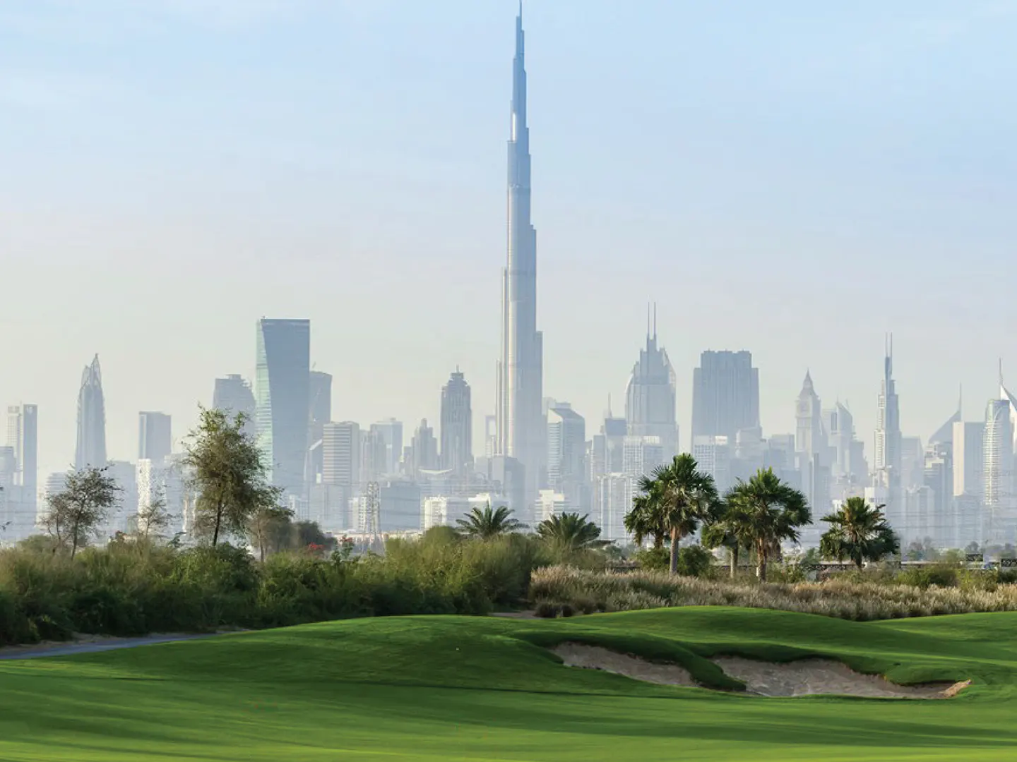 Dubai skyline view from a green golf course with palm trees and a sand bunker in the foreground.