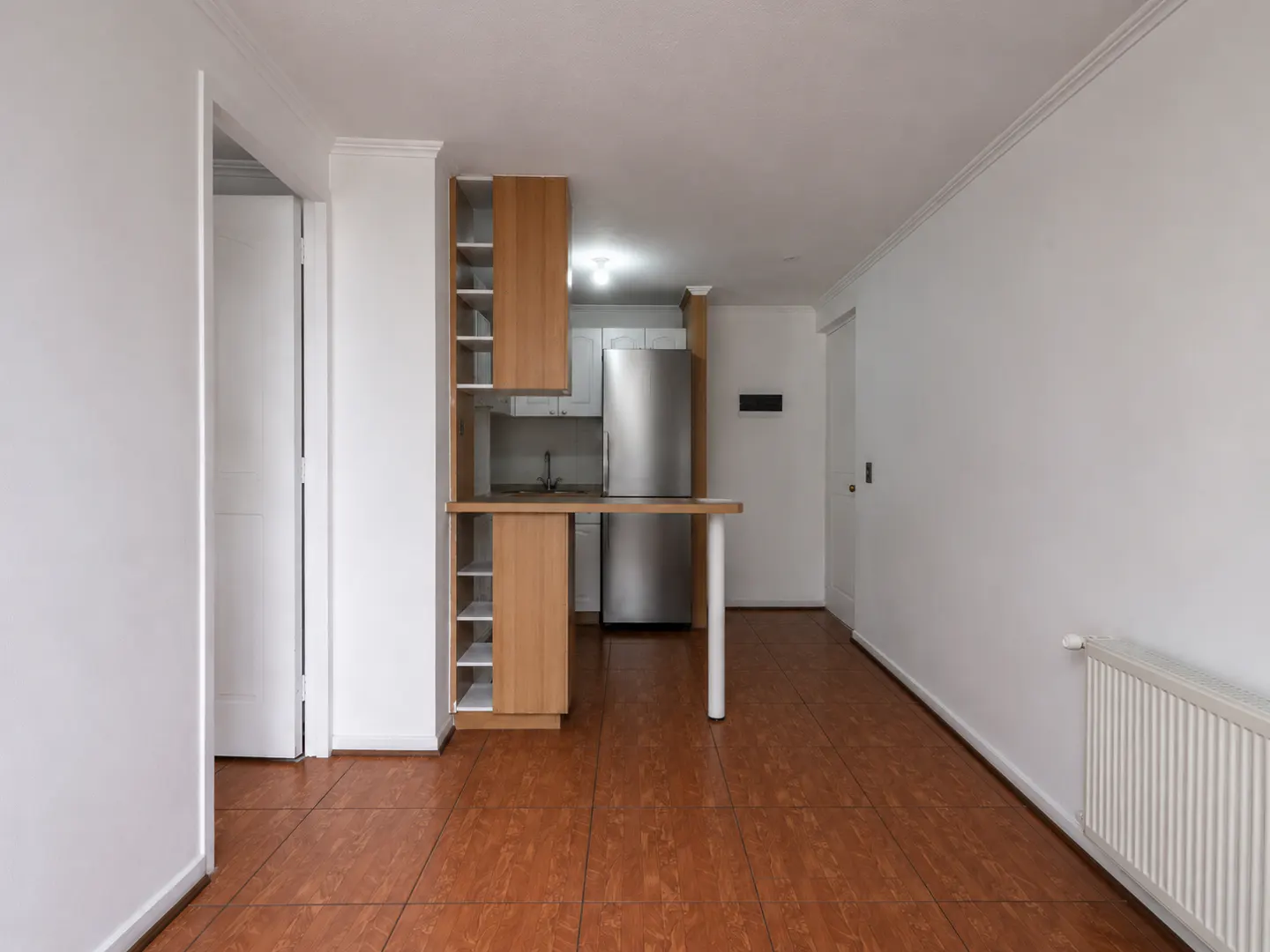 An interior shot of an empty apartment with brown tile floors, white walls, and a small kitchen area.