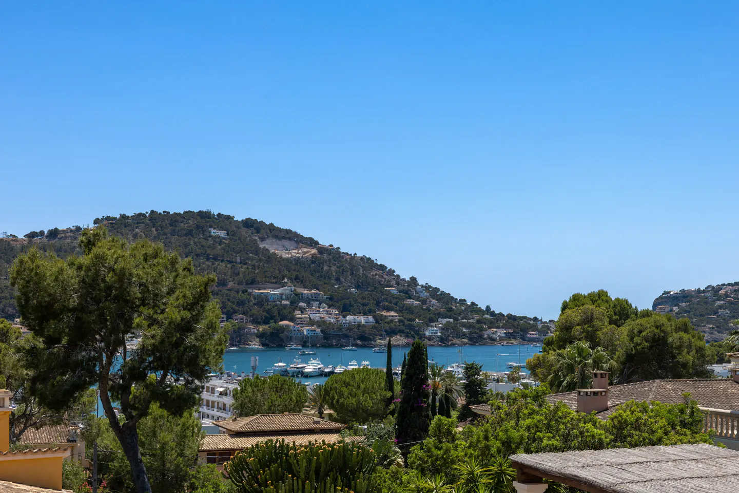 View of a blue bay with yachts, surrounded by lush green hills and Mediterranean-style houses under a clear blue sky.