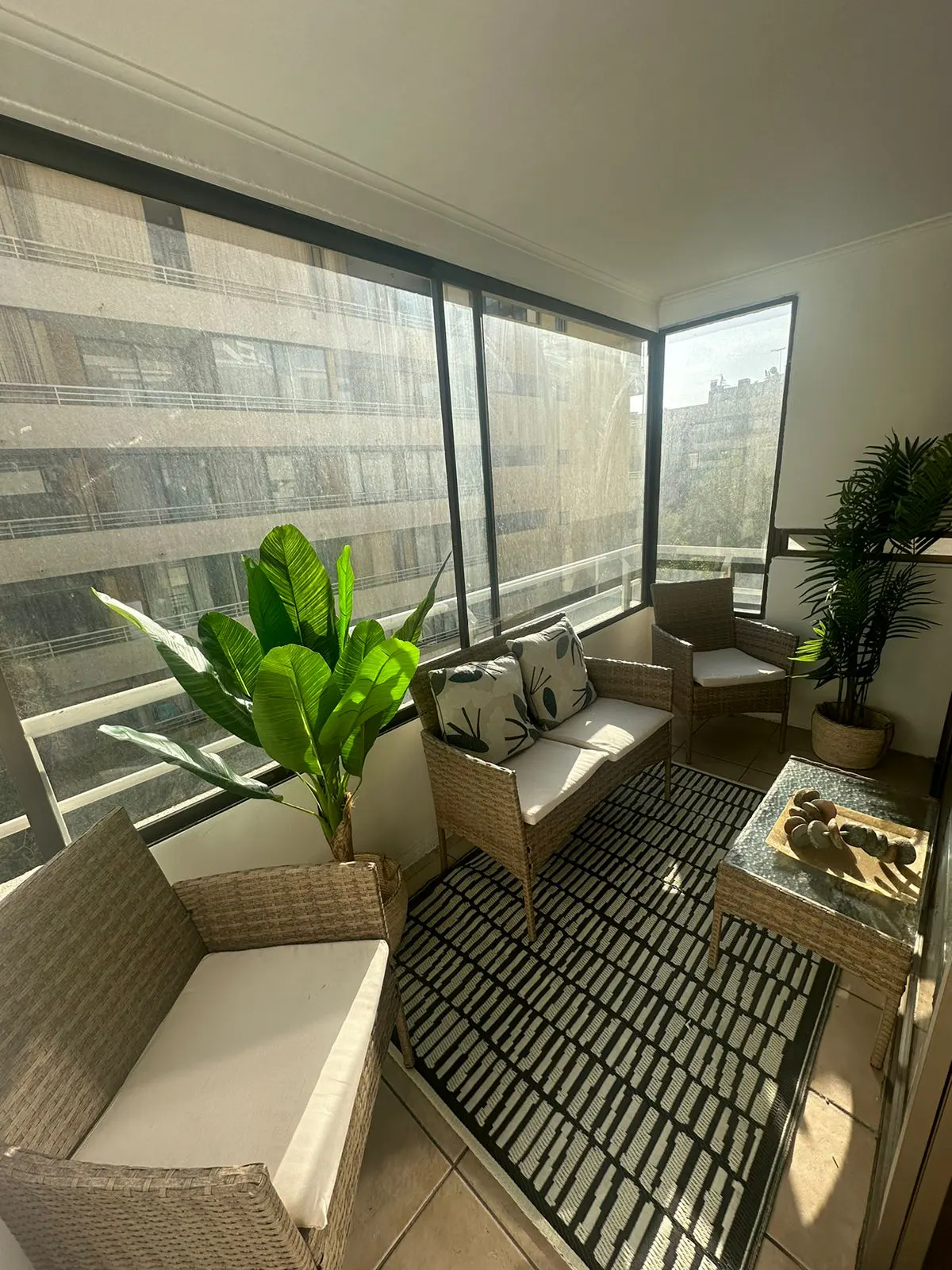 Sunroom with wicker furniture, a black and white rug, and potted plants. Large windows offer a city view.