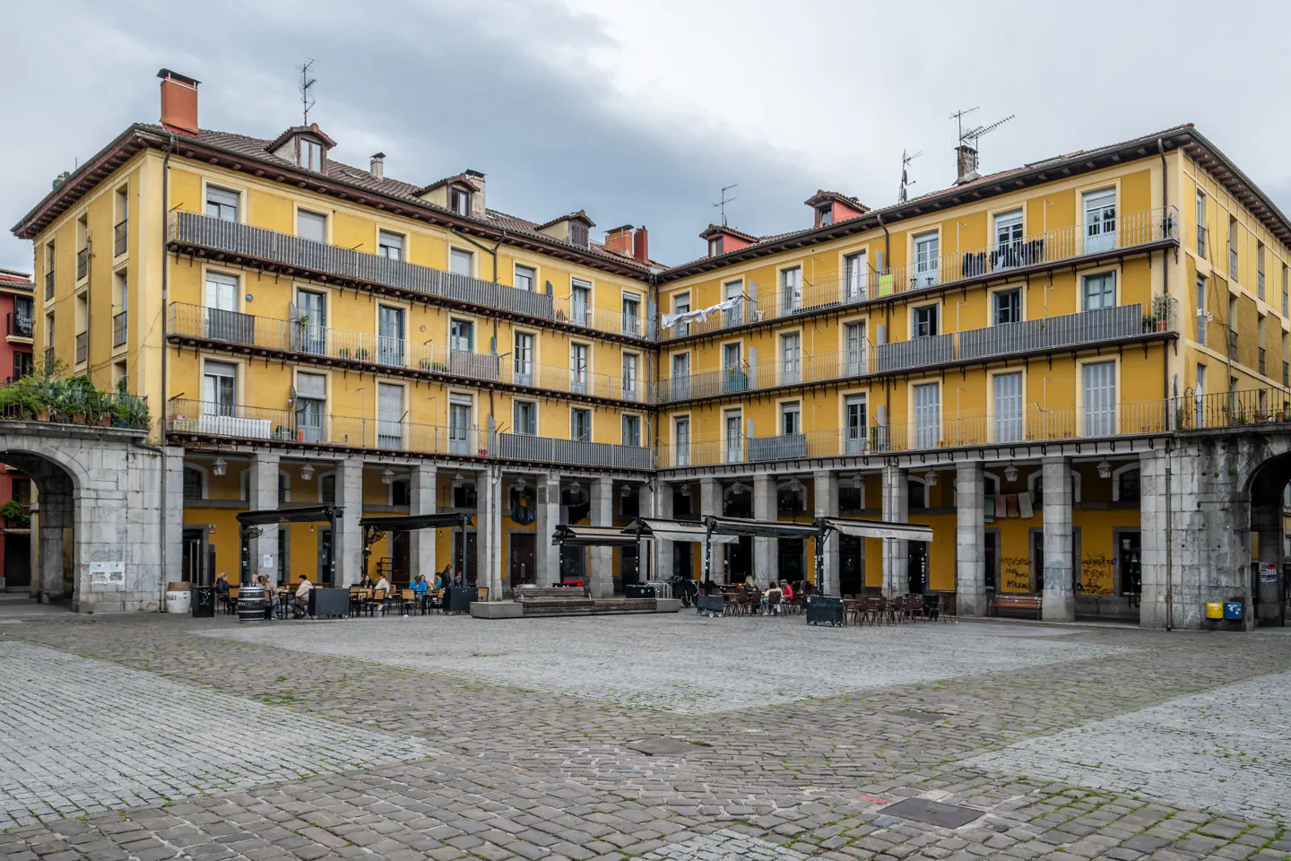 A yellow multi-story building with balconies surrounds a cobblestone plaza featuring outdoor cafe seating under awnings.