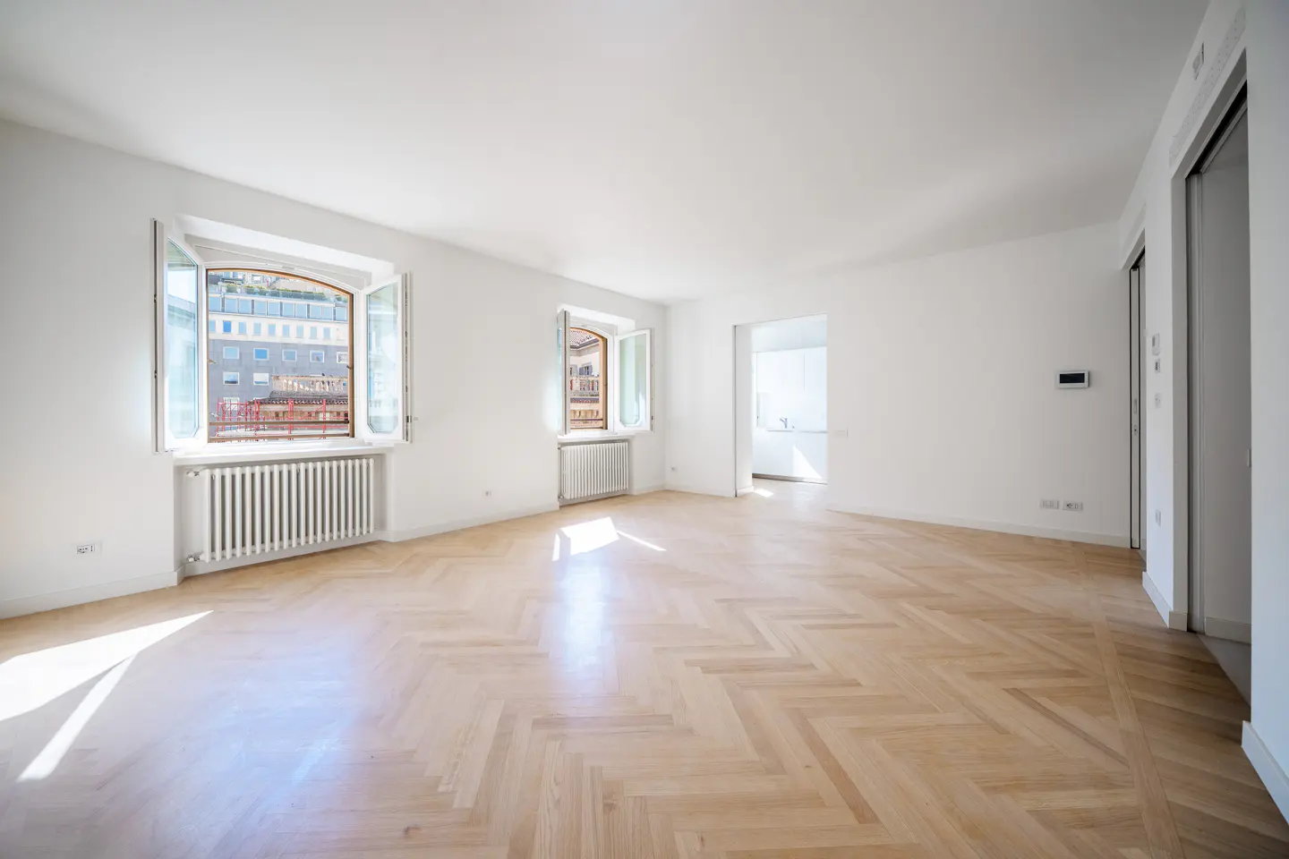 Bright, empty room with herringbone wood floors, white walls, two windows with city views, and a doorway to another room.