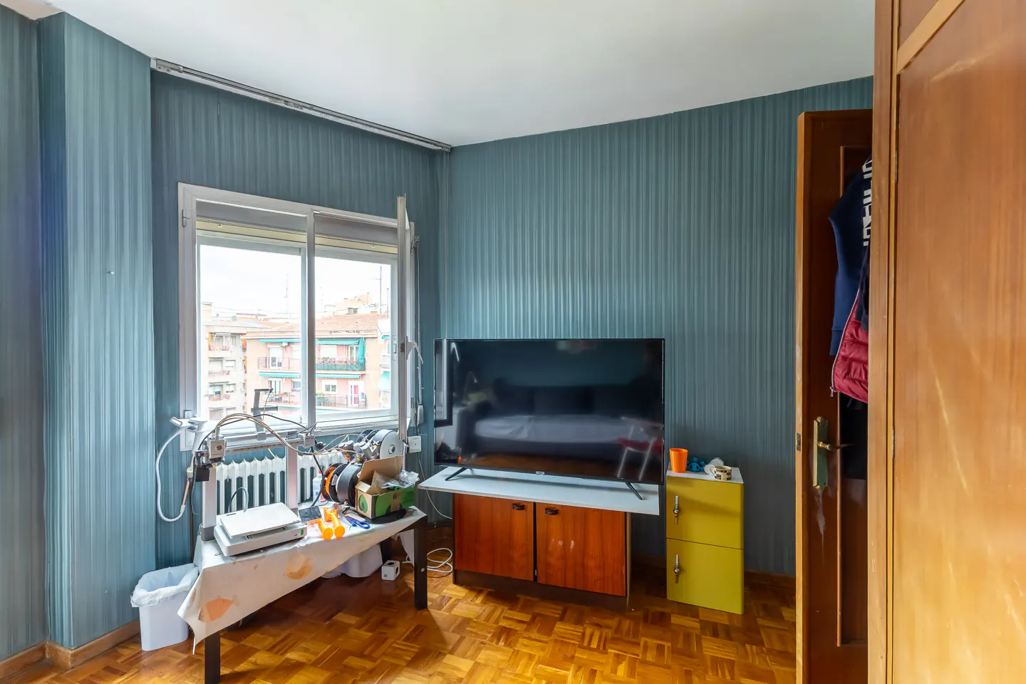 Bedroom with blue striped wallpaper, parquet floor, TV on a cabinet, table with equipment, and open closet.