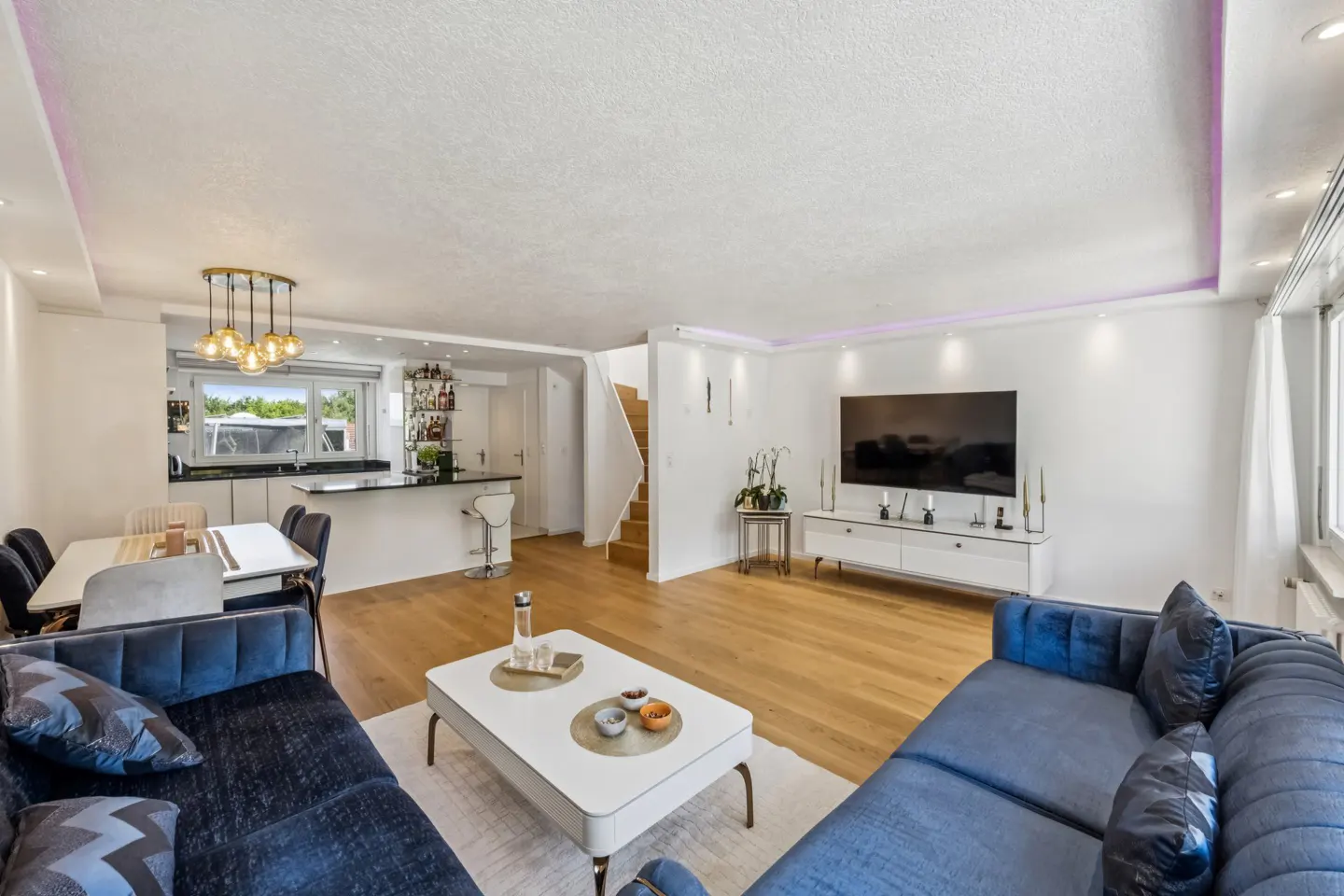 Modern open-plan living room with blue velvet sofas, white coffee table, hardwood floors, and a kitchen bar area.