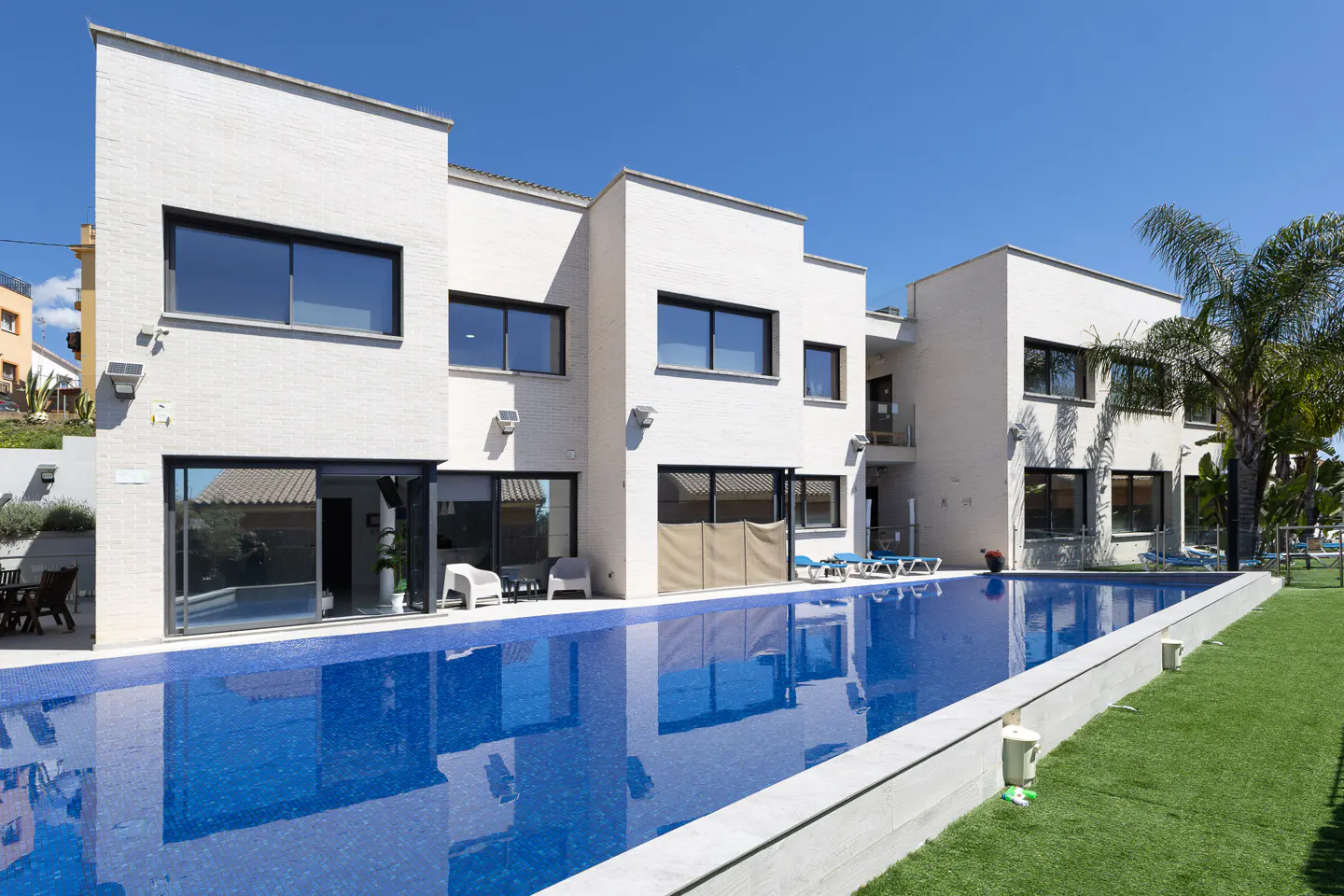 Modern white house with a blue tiled pool, green lawn, and palm trees under a clear blue sky.
