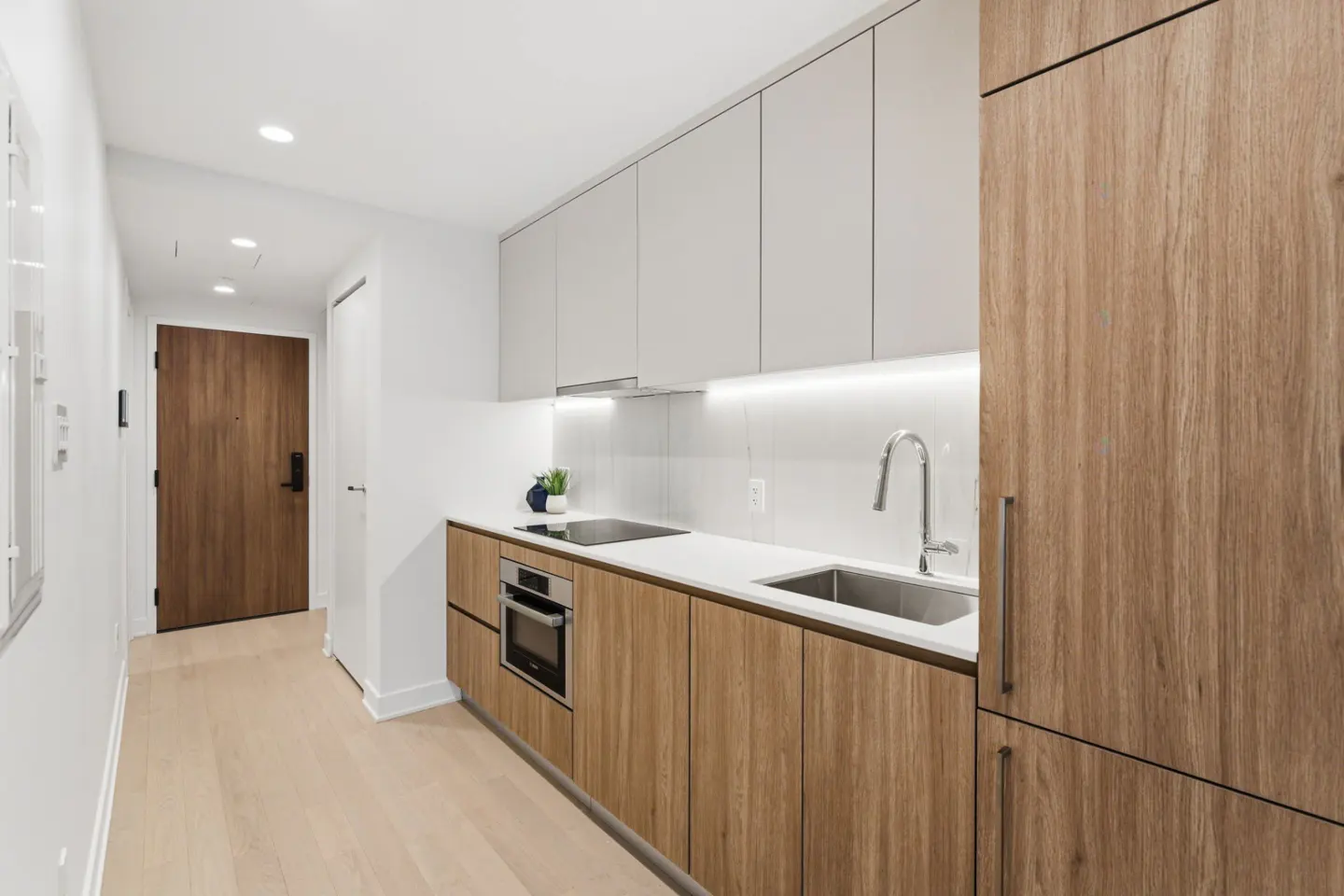 A modern kitchen with light wood cabinets, white countertops, and stainless steel appliances. A wood door is visible in the hallway.