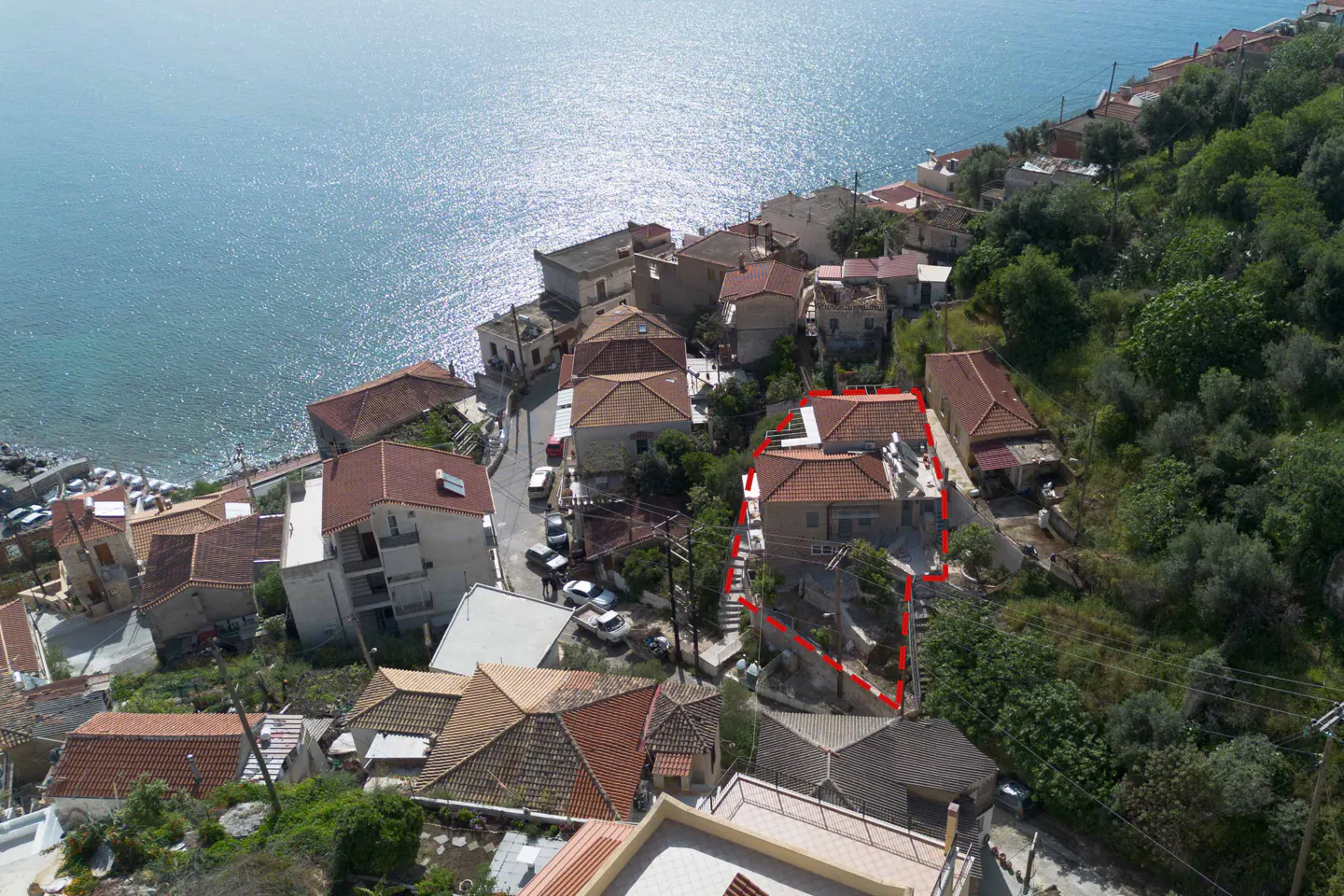 Aerial view of a house with a red tile roof, outlined in red, near the sea and a hillside with trees.