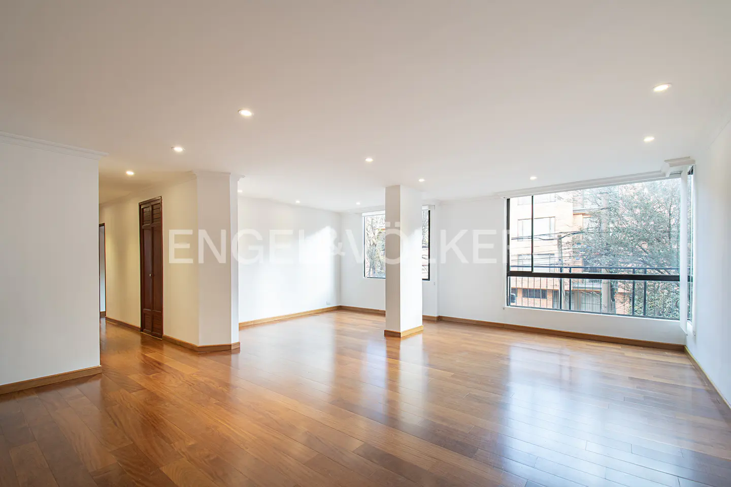 Bright, empty room with hardwood floors, white walls, and large windows showing trees and buildings outside.