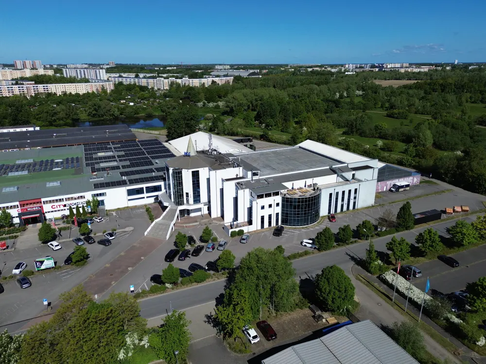 Aerial view of a large, white commercial building with a parking lot, surrounded by green trees and distant buildings.