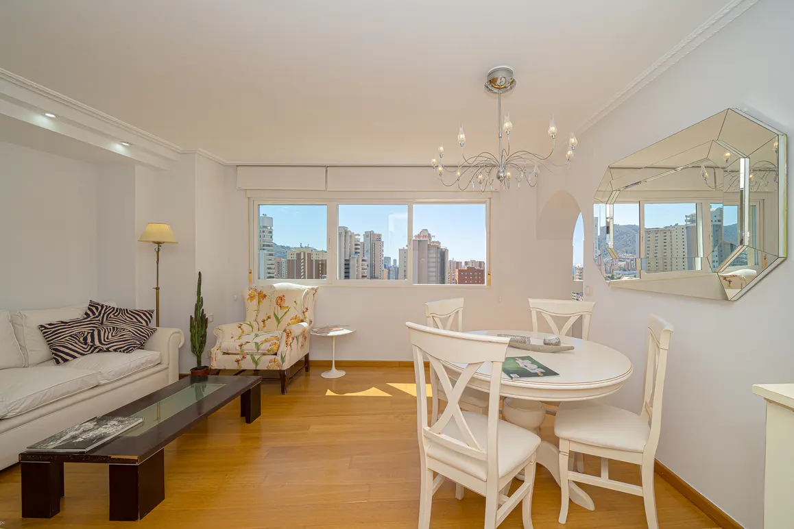 Bright living room with white sofa, floral armchair, and dining table. Large windows offer a city view. A chandelier hangs above.