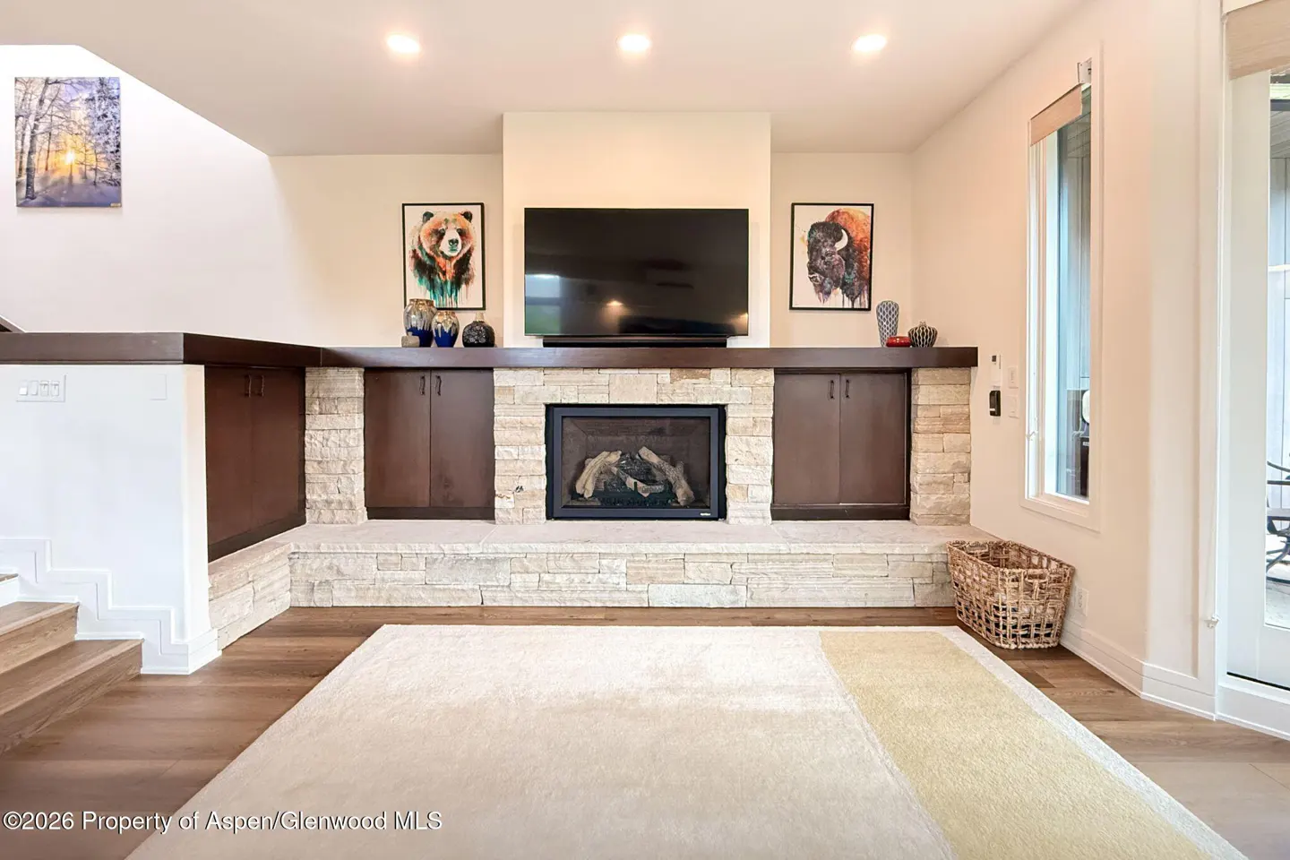 Living room with a stone fireplace, flat-screen TV, dark wood cabinets, bear and bison art, and a large area rug.
