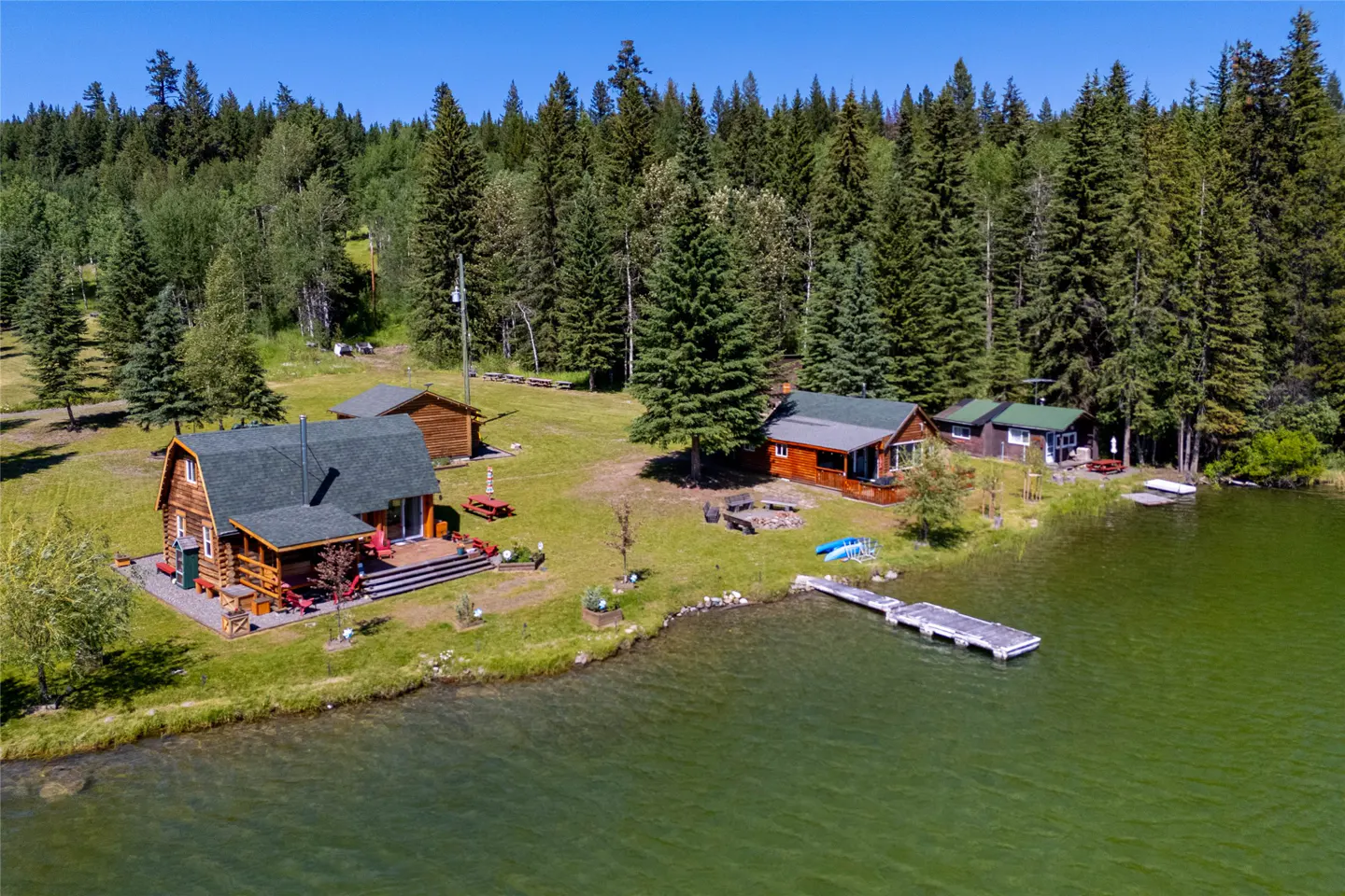 Aerial view of log cabins with green roofs on a lakefront property surrounded by tall green trees. A dock extends into the green water.