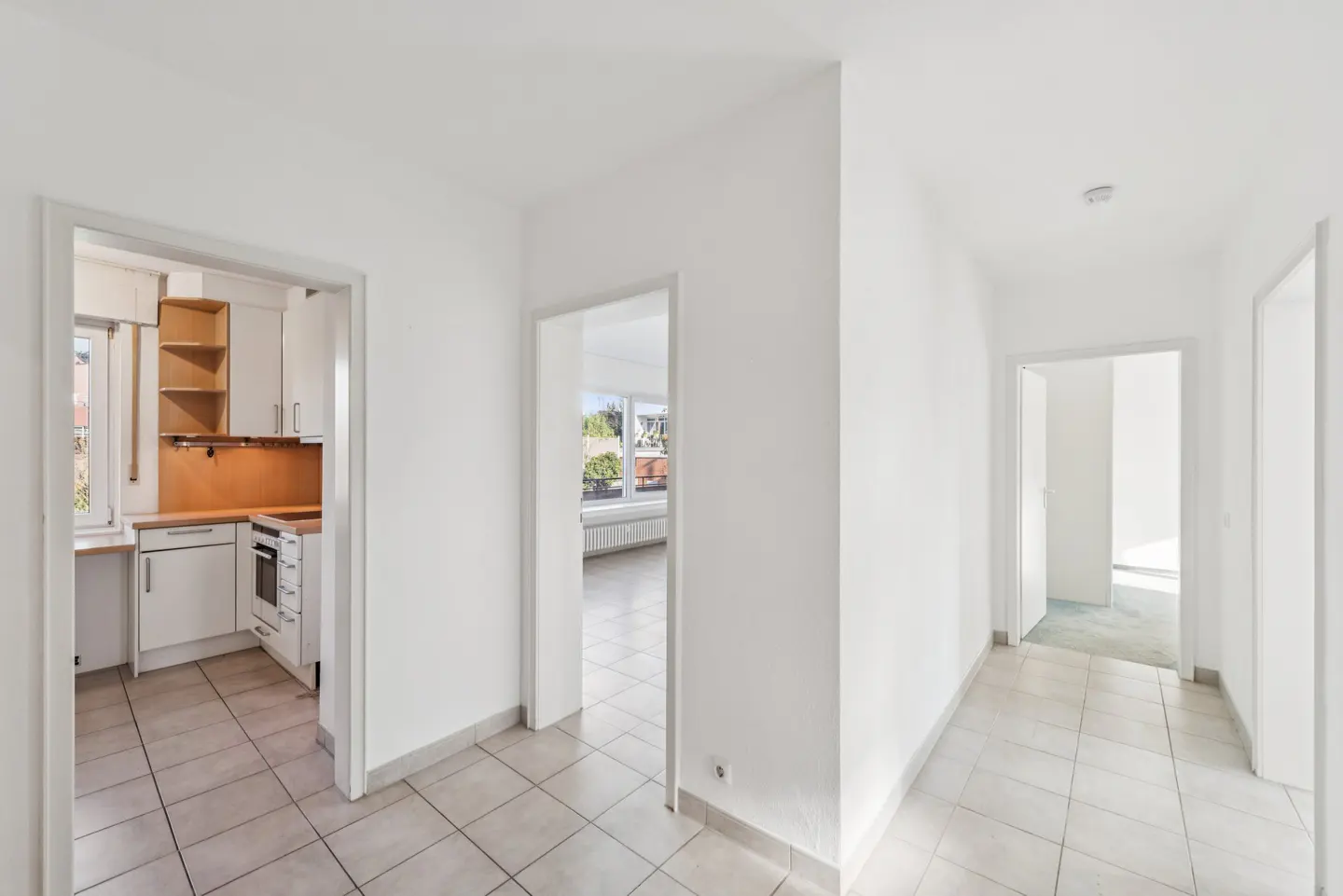 Hallway view of a bright, white apartment with tile floors, leading to a kitchen and living room.