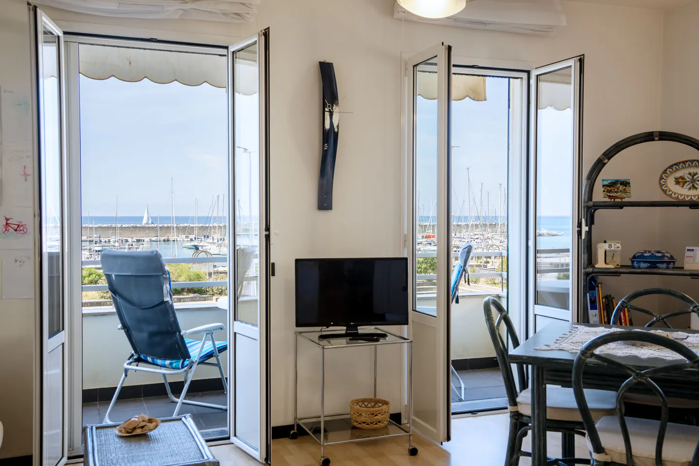 Living room with open doors to a balcony overlooking a marina with sailboats, a TV on a stand, and a dining area.