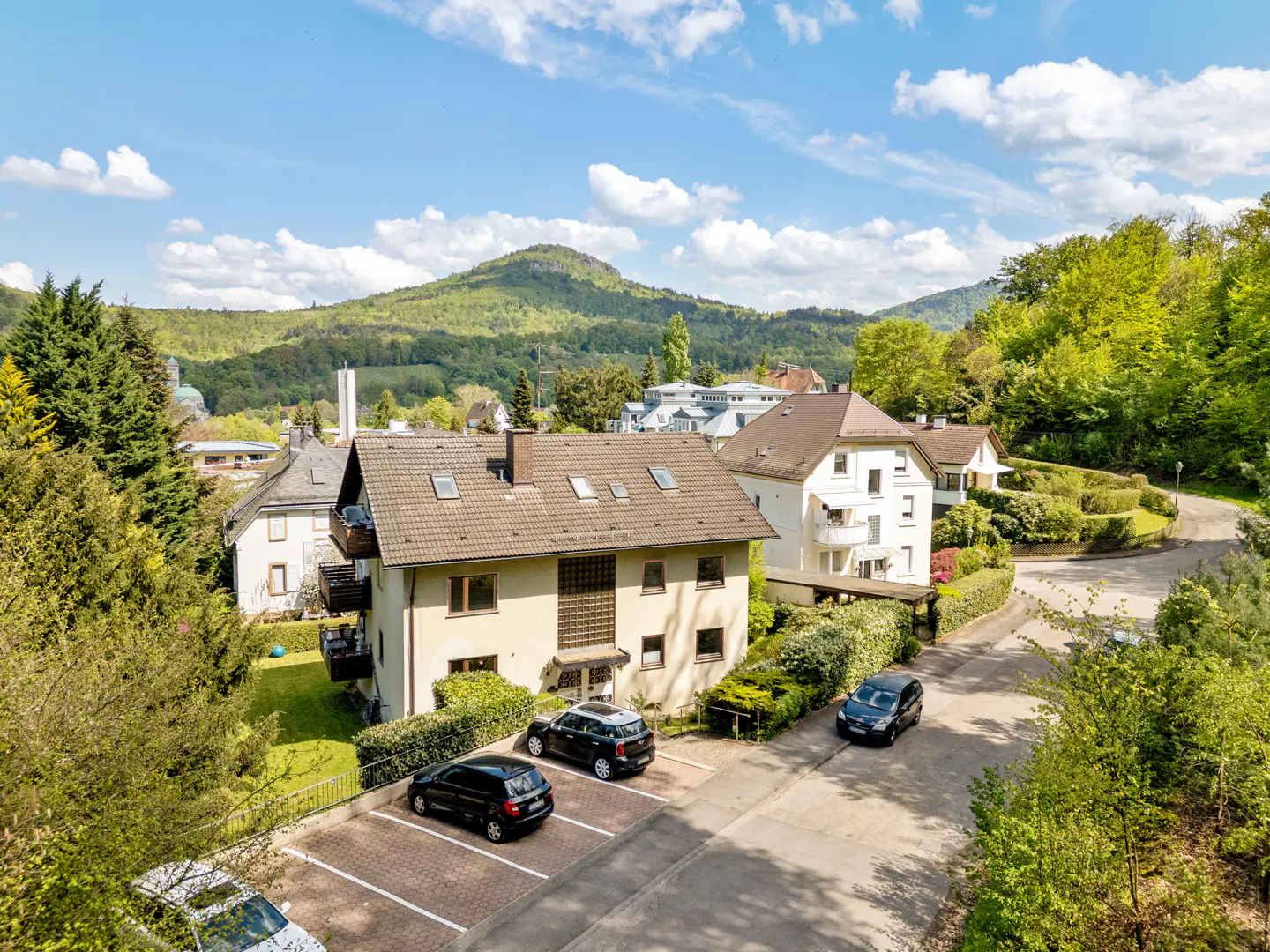View of a beige house with a brown roof, cars parked in front, and a green, mountainous background.