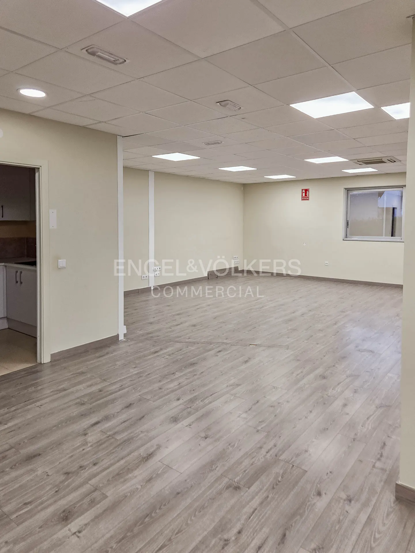 Bright, empty commercial space with gray wood floors and white walls. A kitchen area is visible through a doorway. Engel & Volkers logo on the wall.