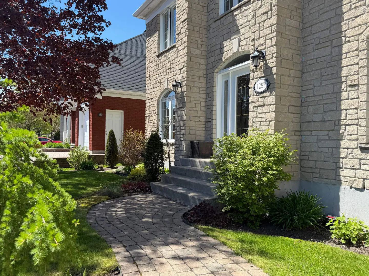 Exterior of a two-story stone house with a white door and a brick walkway leading to the entrance. Green lawn and trees surround the house.