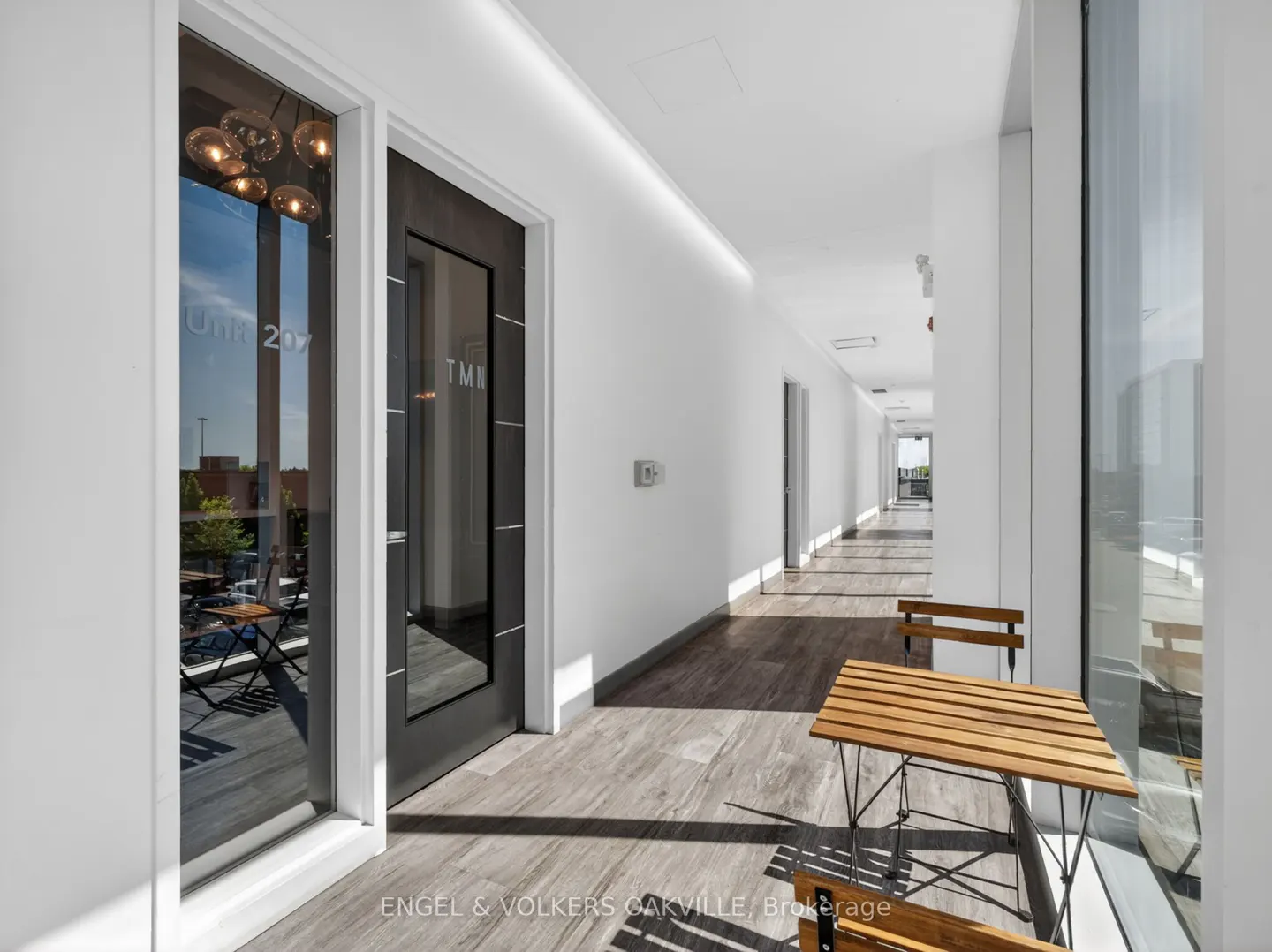 Bright hallway with white walls, gray wood floors, and a wooden table and chairs. Glass doors lead to outdoor seating.