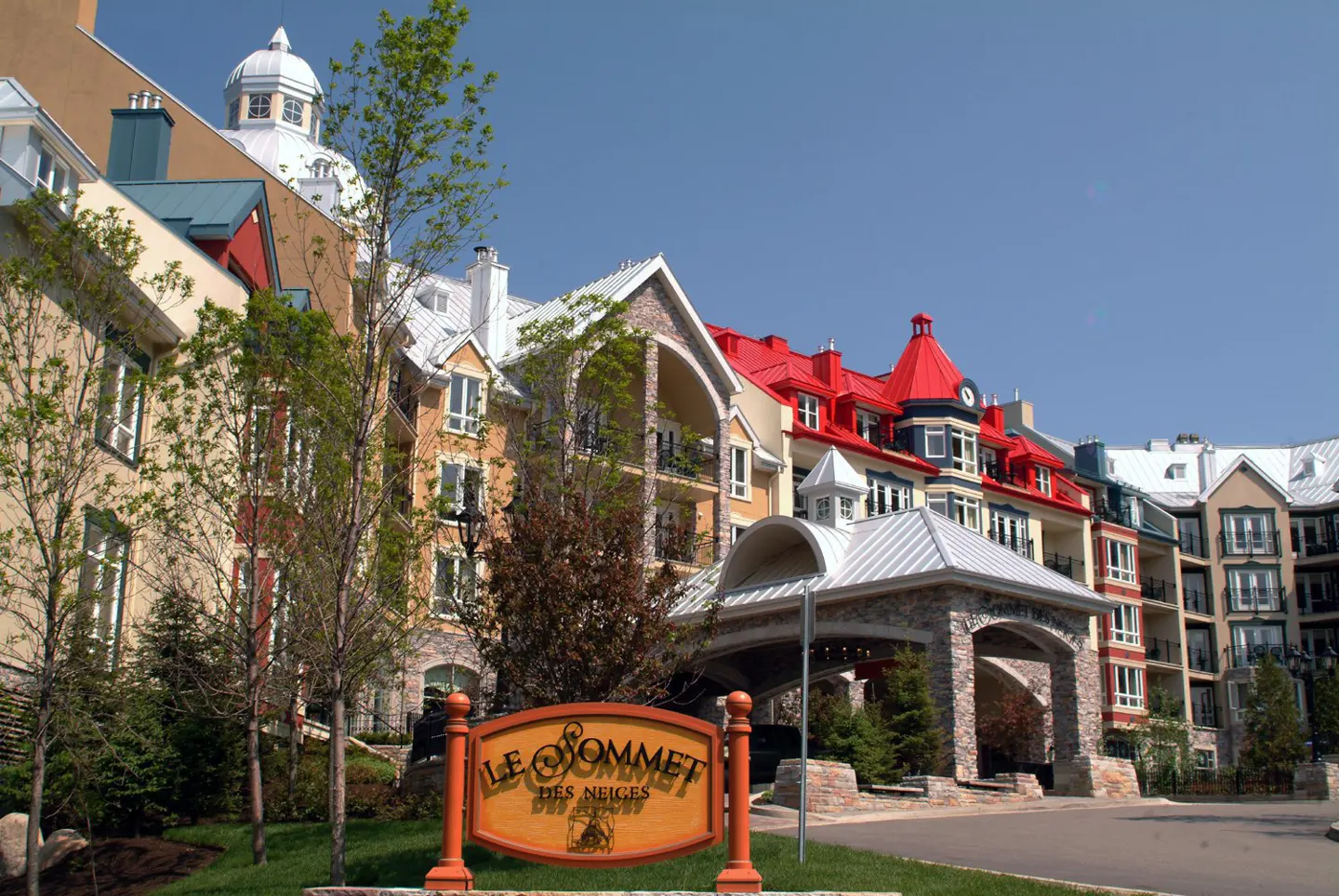 Exterior view of Le Sommet Des Neiges, a multi-story resort with colorful roofs and a stone entrance. A wooden sign is in the foreground.