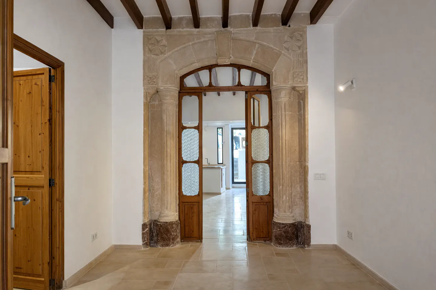 Interior view of a room with white walls, beige tile floor, and an arched doorway with wooden doors and stone frame.
