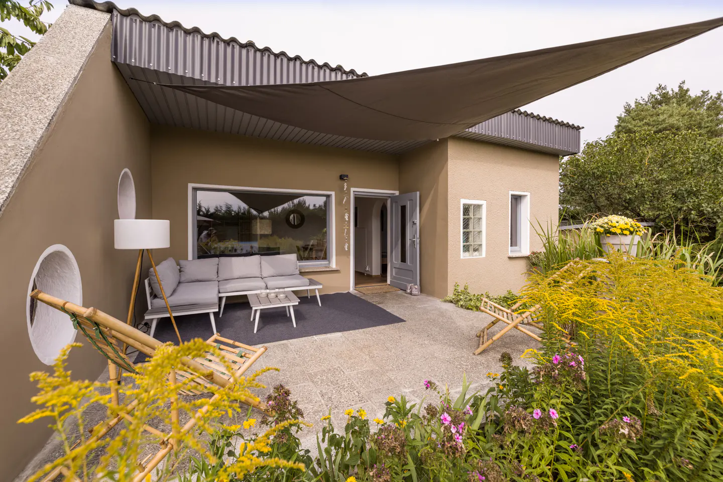 Tan house with a patio featuring a gray sofa, table, and sunshade. Flowers and greenery surround the patio.