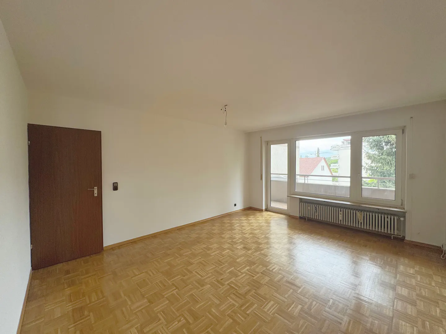 Empty room with parquet floor, white walls, and a large window with a view of houses and trees. A brown door is on the left.