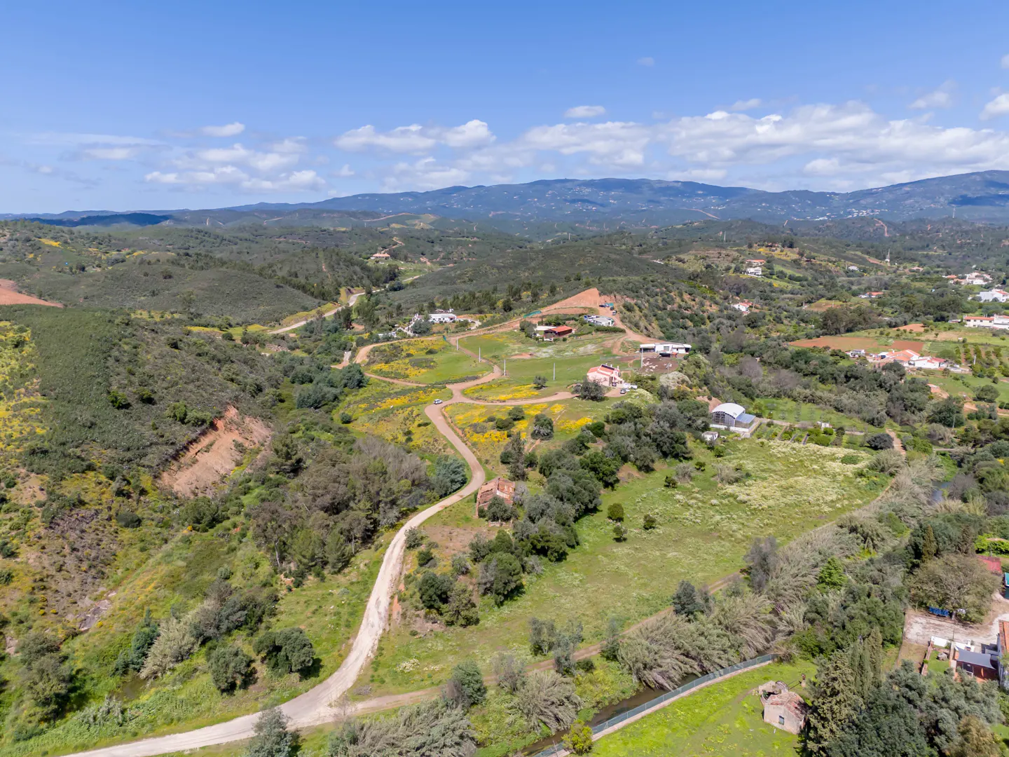 Aerial view of a rural landscape with green hills, scattered houses, and a winding dirt road under a blue sky.