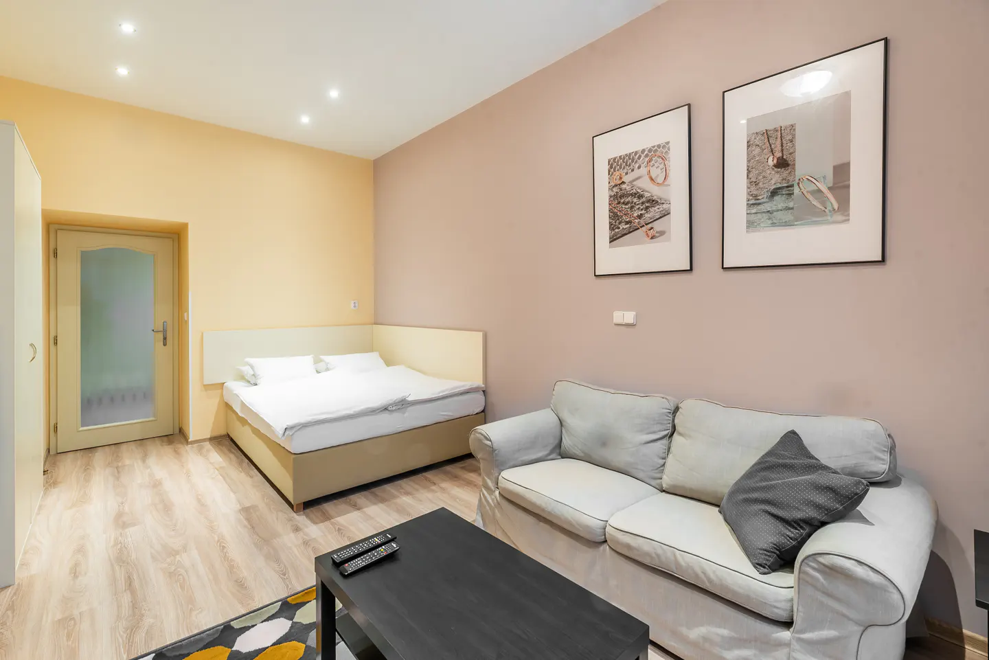 A studio apartment with a bed, a light grey sofa, a black coffee table, and two framed pictures on a beige wall.