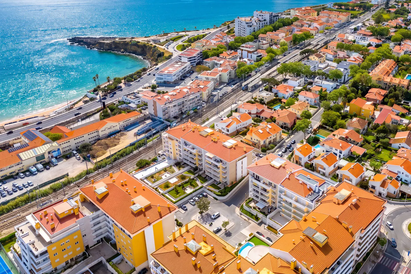 Aerial view of a coastal town with orange-roofed buildings, a sandy beach, and turquoise ocean water.