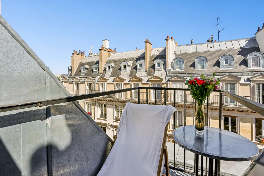 Balcony view with white chair, table with flowers, and Parisian buildings under a blue sky.