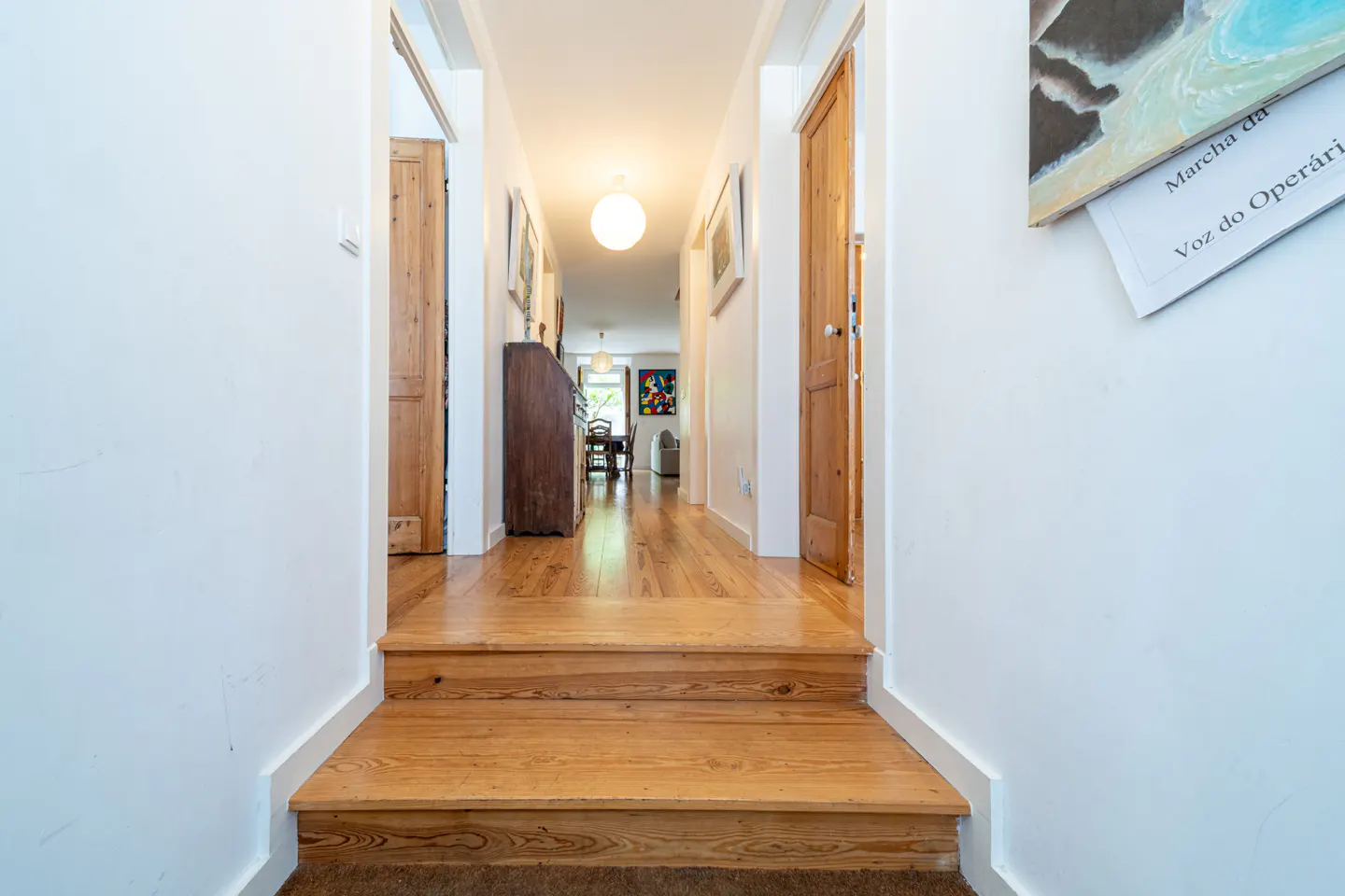 Interior view of a hallway with wooden floors and white walls, leading to a dining area. Two wooden doors are visible on either side of the hallway.