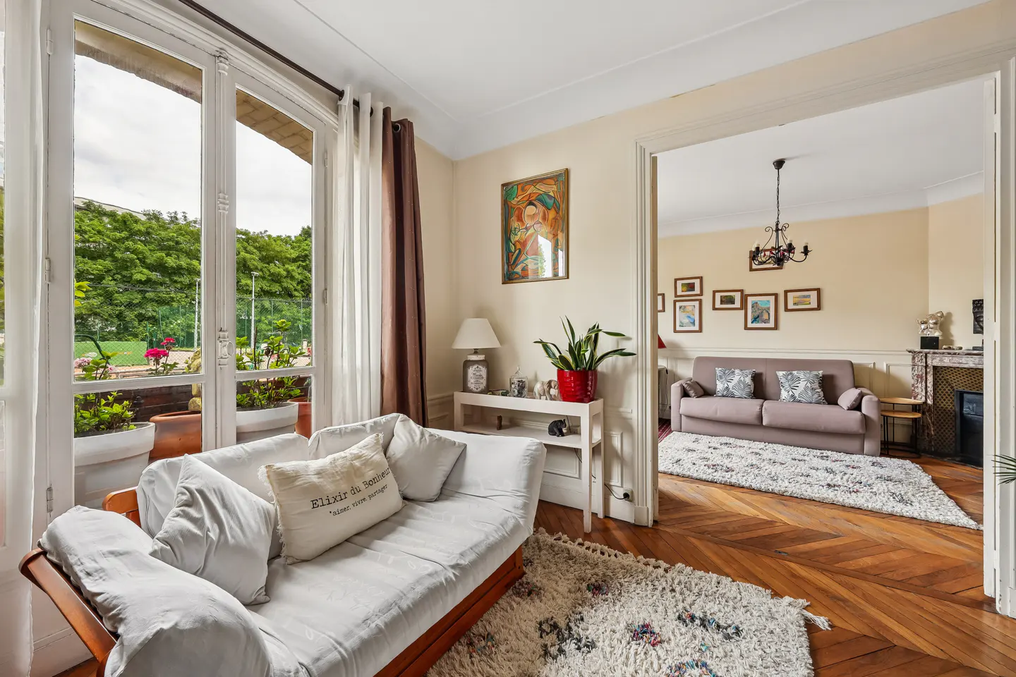 Living room with white sofa, wood floors, and view to a garden through a glass door. Another room is visible through a doorway.