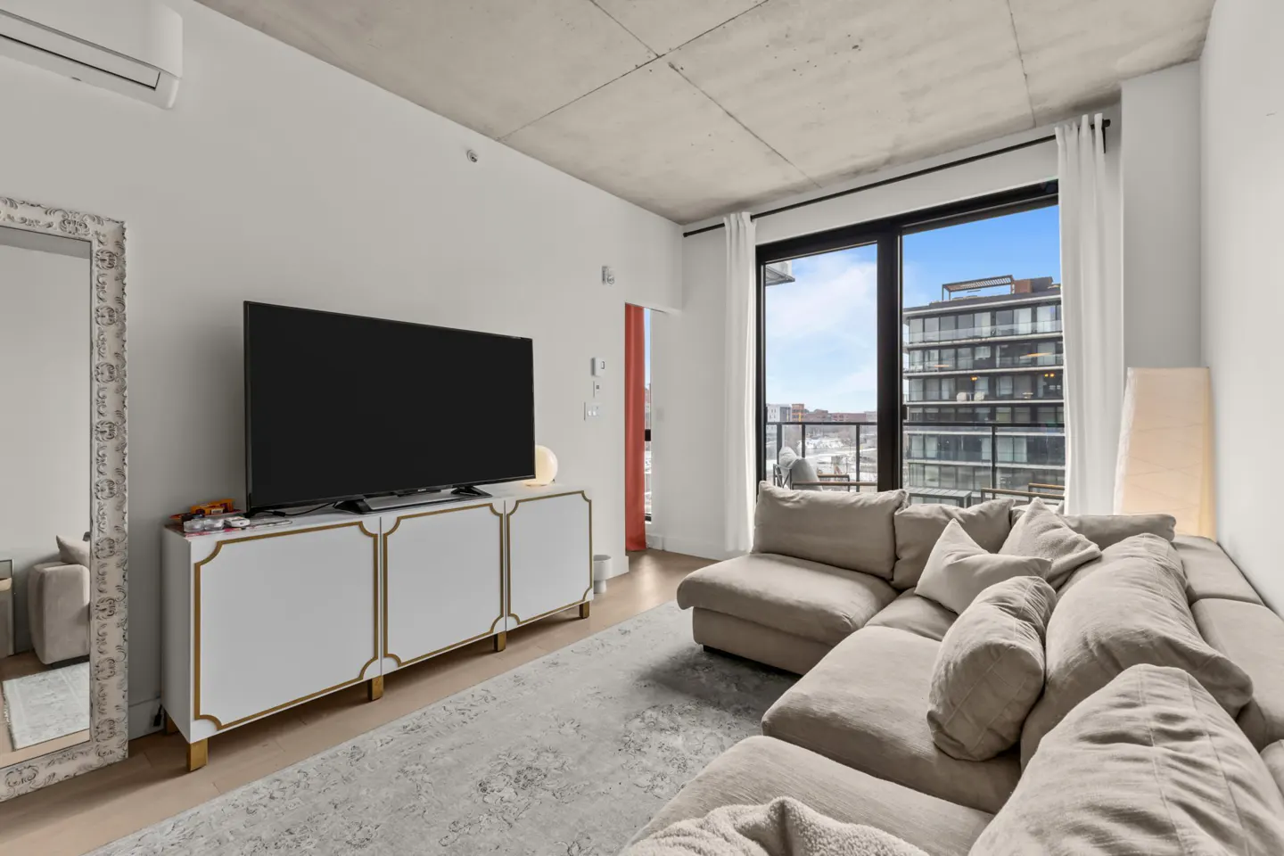 Living room with a large beige sectional sofa, a white and gold media console with a TV, and a large window with city views.