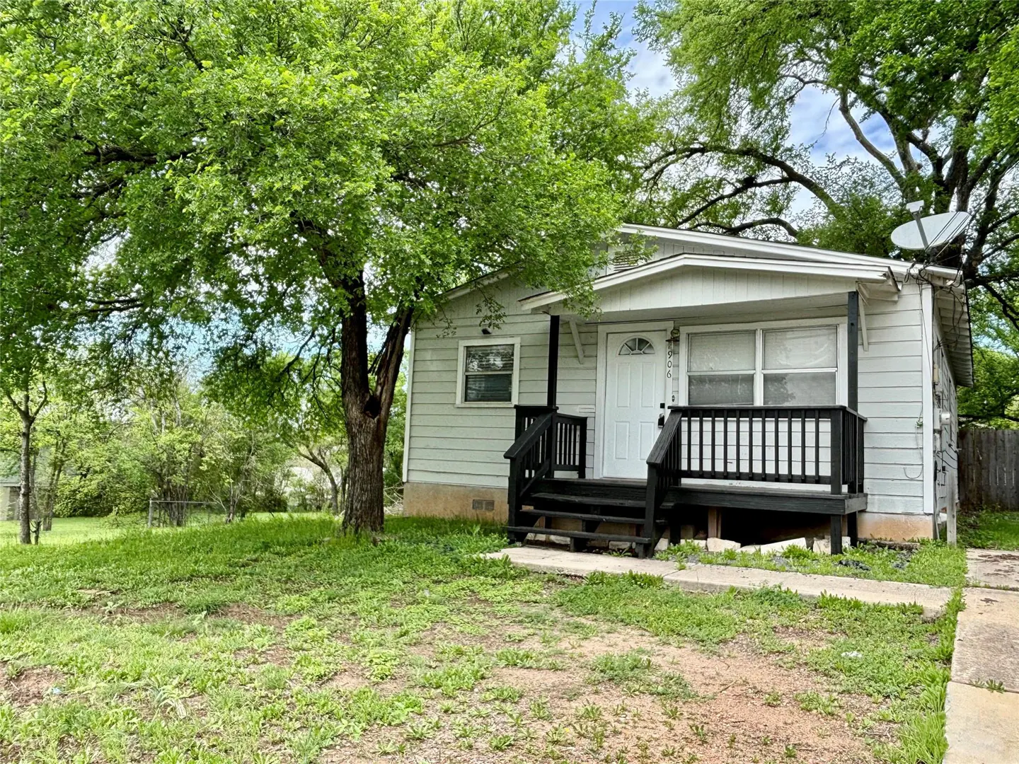 A light gray, one-story house with a white door and black porch railing sits on a green lawn with a large tree.