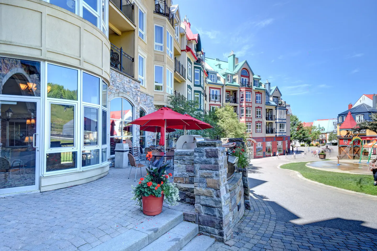 Outdoor patio with red umbrellas, stone planters, and colorful buildings in the background on a sunny day.
