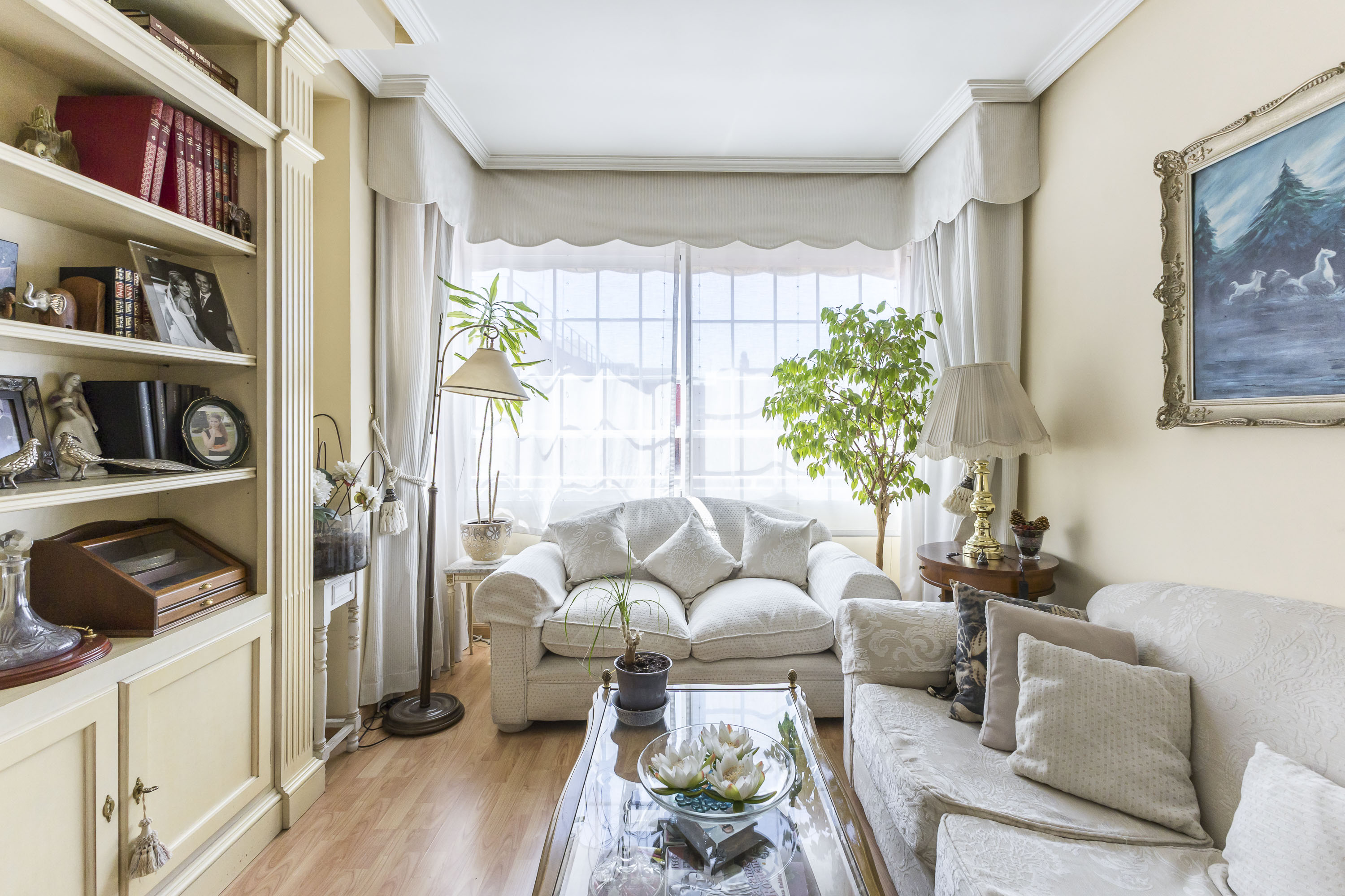 Cozy living room with cream sofas, a glass coffee table, built-in bookshelves, and a large window with white curtains. [AI-generated text]