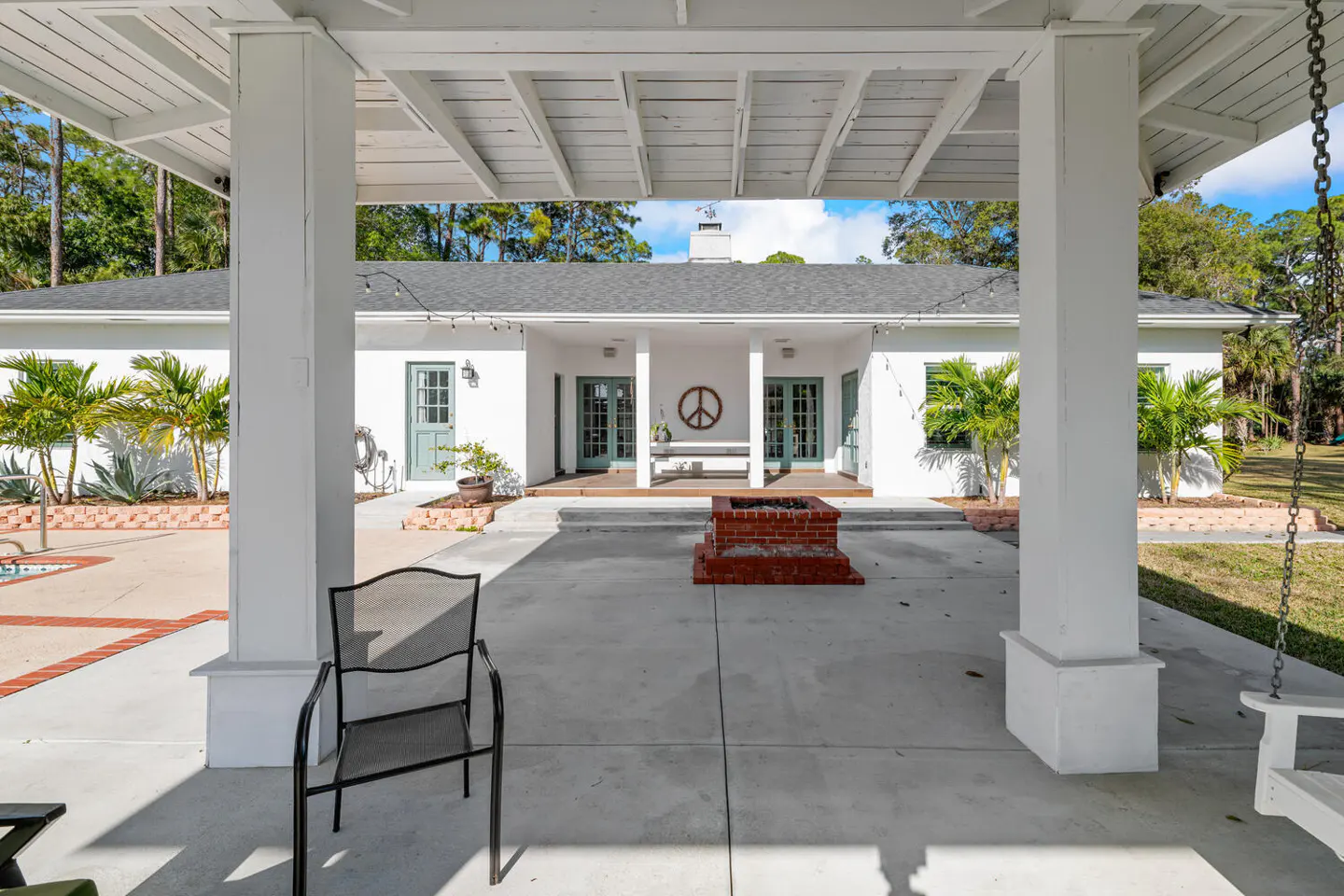 View of a white house with a gray roof, seen from a covered patio with white pillars and a concrete floor. A brick fire pit sits in the yard.