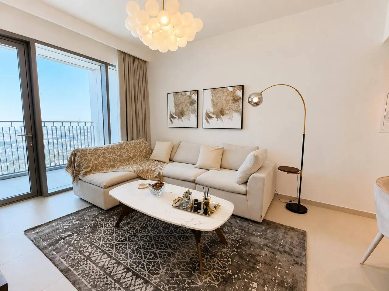 Bright living room with a beige sectional sofa, marble coffee table, and patterned rug. A gold floor lamp and city view add elegance.