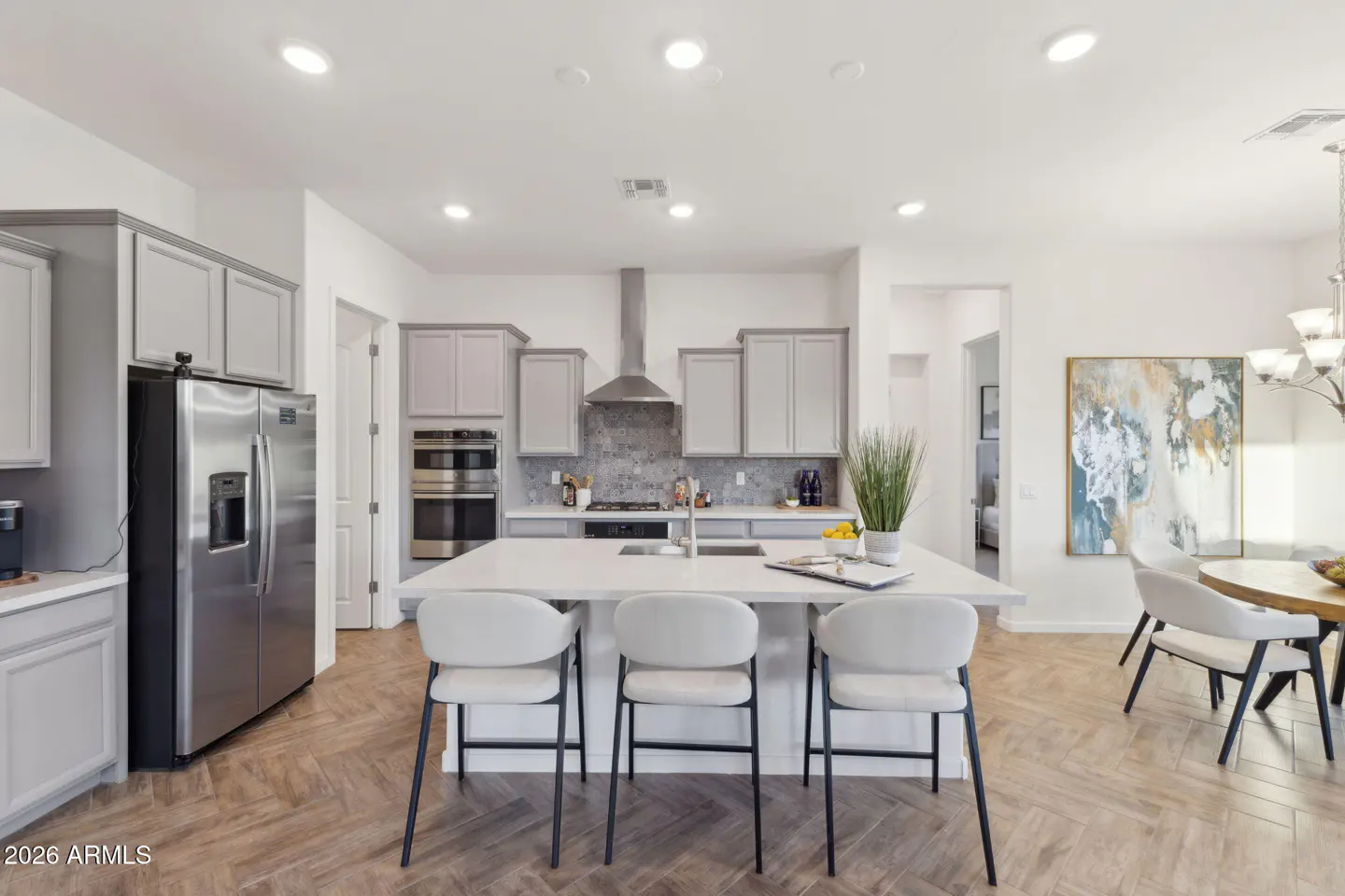 A modern kitchen with gray cabinets, stainless steel appliances, and a white island with three chairs. A dining table is visible in the background.