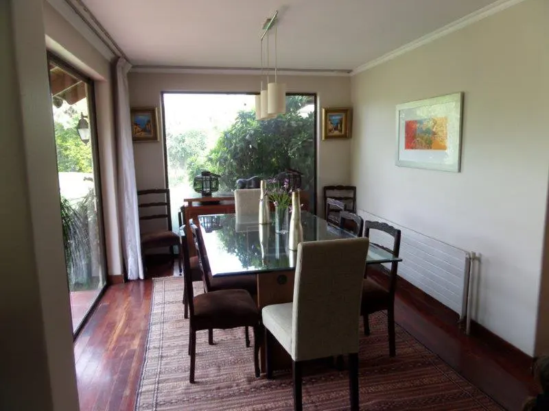 Dining room with a glass-topped table, brown chairs, and a beige rug on a dark wood floor. A large window overlooks green foliage.