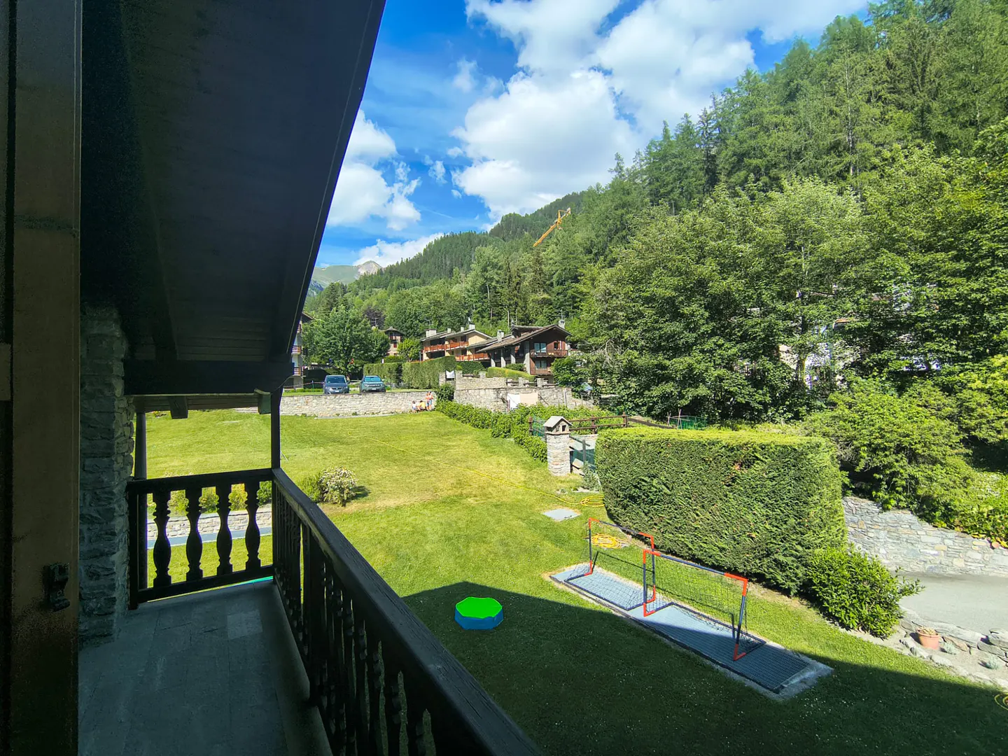 View from a wooden balcony overlooking a lush green lawn with a small soccer goal, a play pool, and mountain scenery.