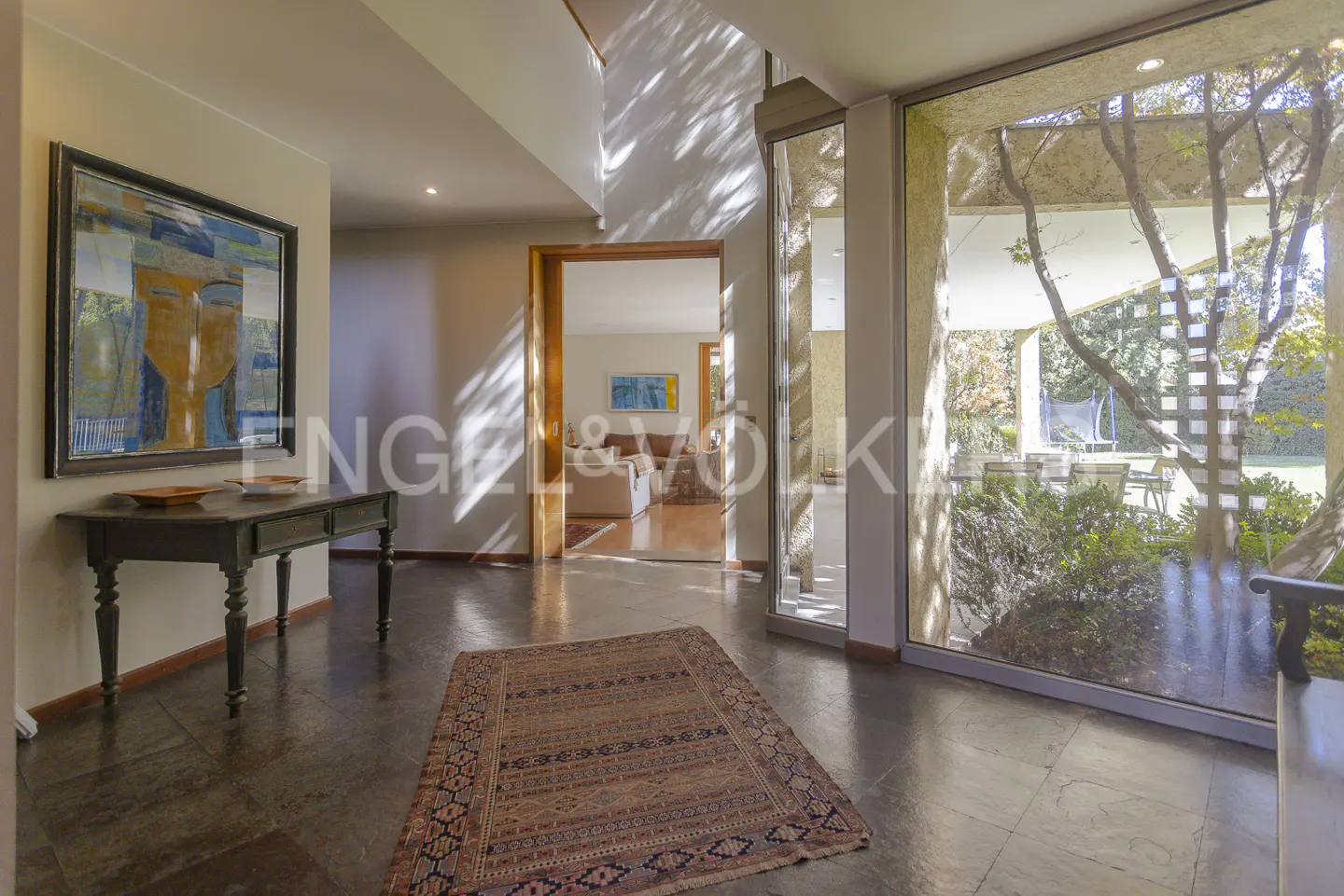 A home's foyer with a dark wood floor, a patterned rug, a table with a painting above it, and a large window.