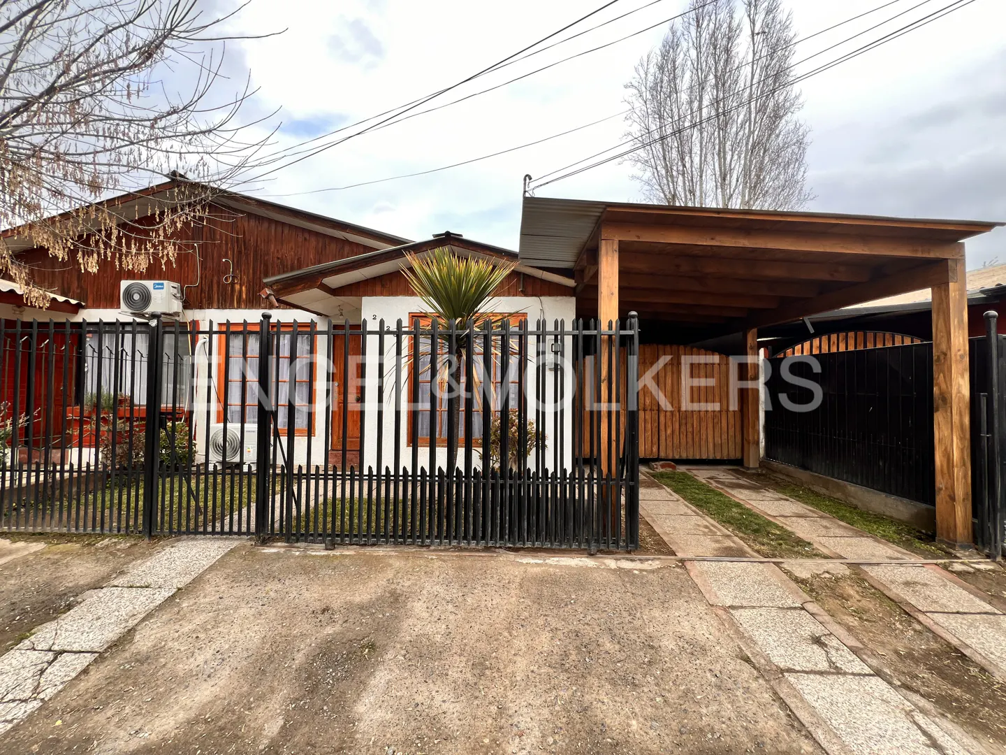 A single-story house with a black metal fence, a wooden carport, and a concrete driveway.